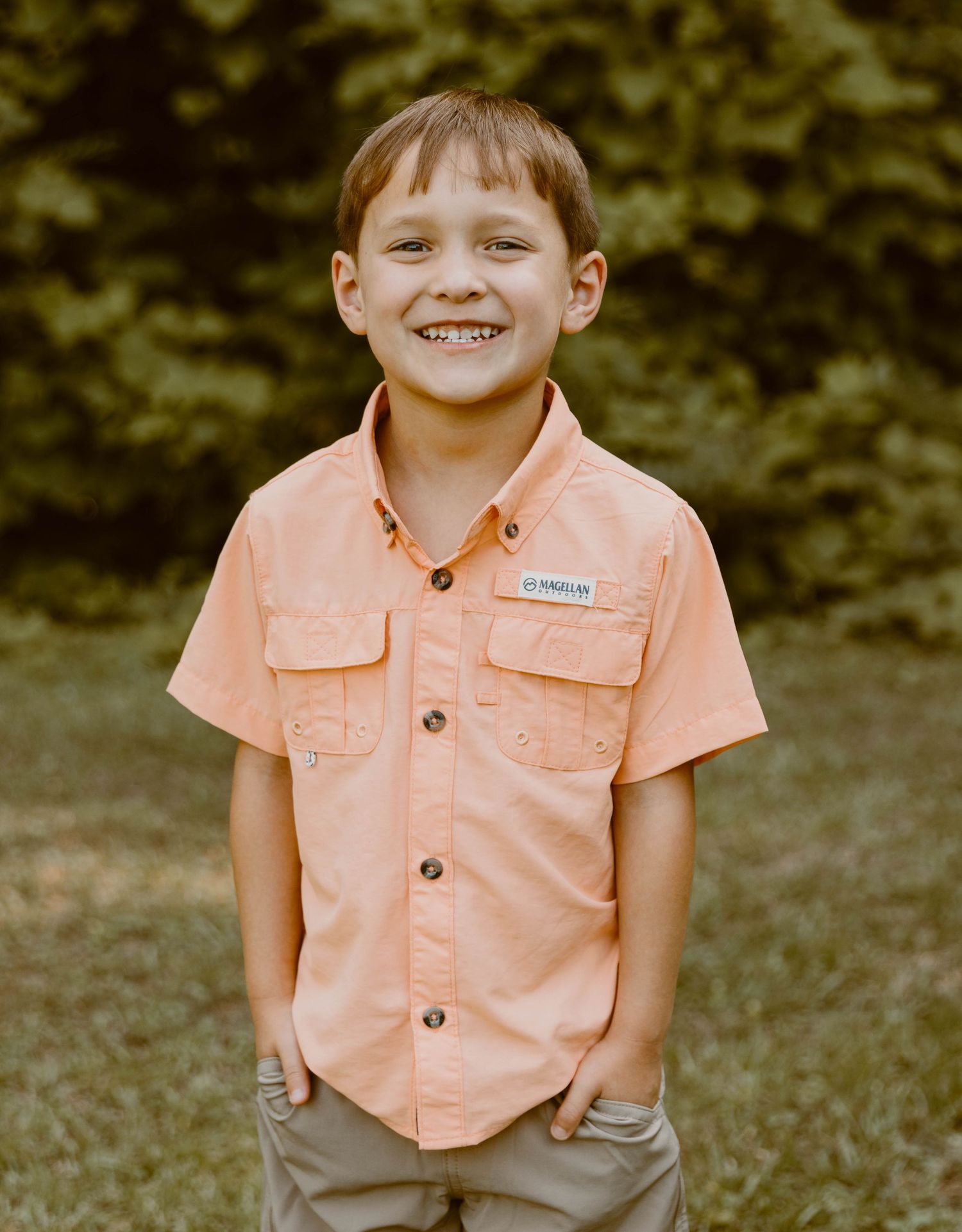 Child in peach button-up shirt and khaki shorts sitting on grass outdoors.