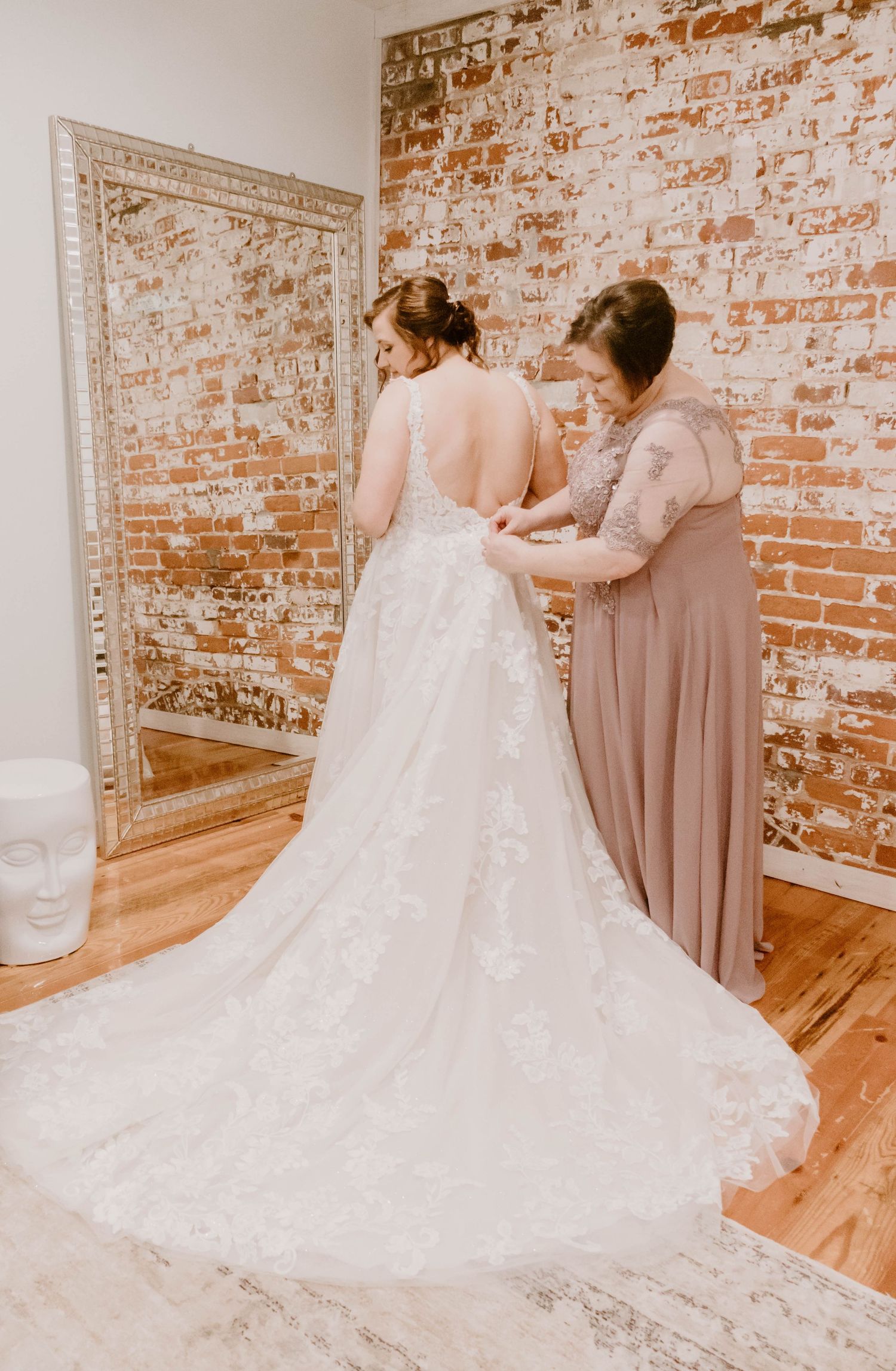 Elegant white wedding dress with lace details and long train being adjusted against exposed brick wall.