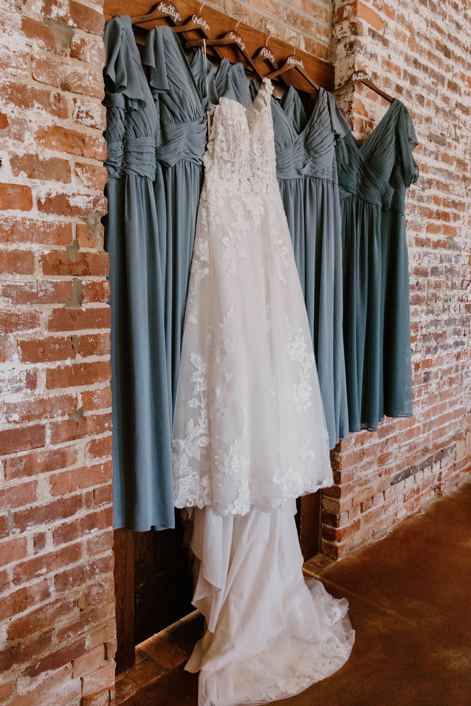 Bridesmaid dresses in slate blue hang alongside a white wedding gown against an exposed brick wall.