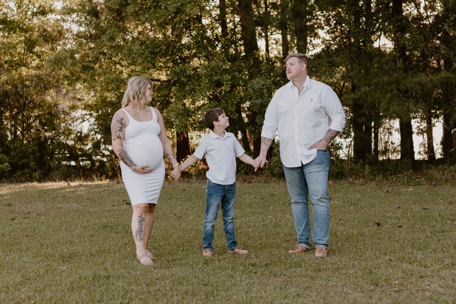 A family walks together holding hands in a sunlit grassy field during a maternity photo session.