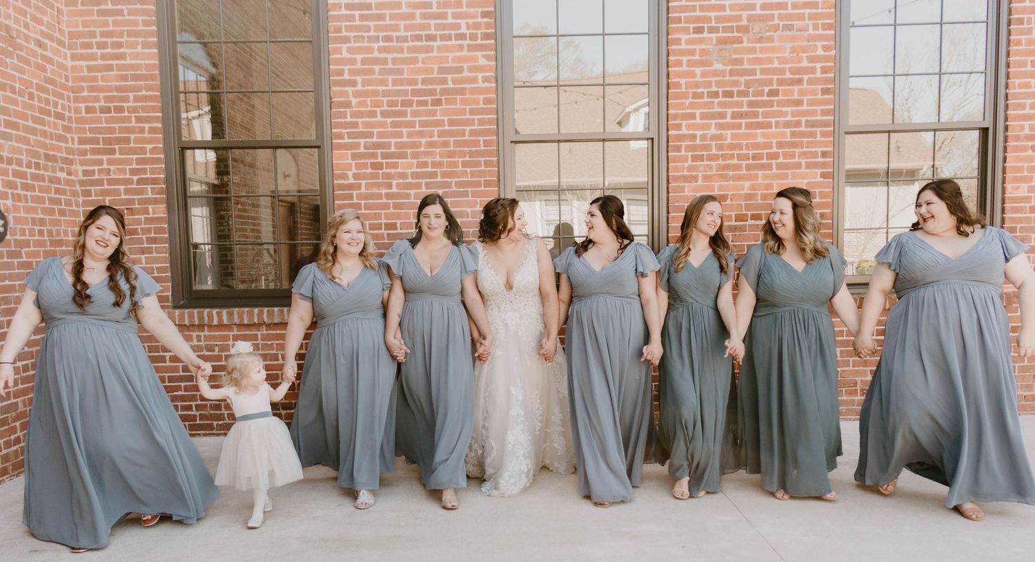 Wedding party in blue dresses creating fun poses against industrial brick wall.