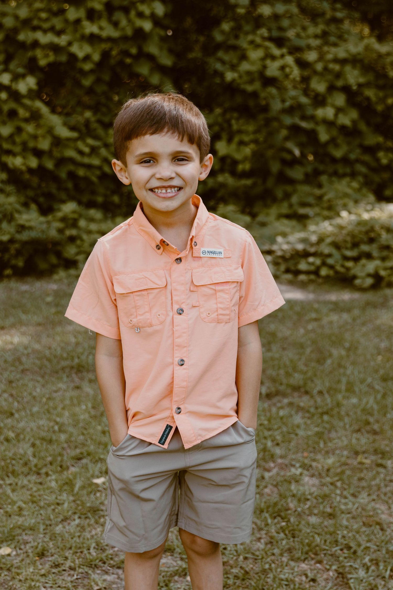 Child wearing peach colored shirt and gray shorts posing on grass.