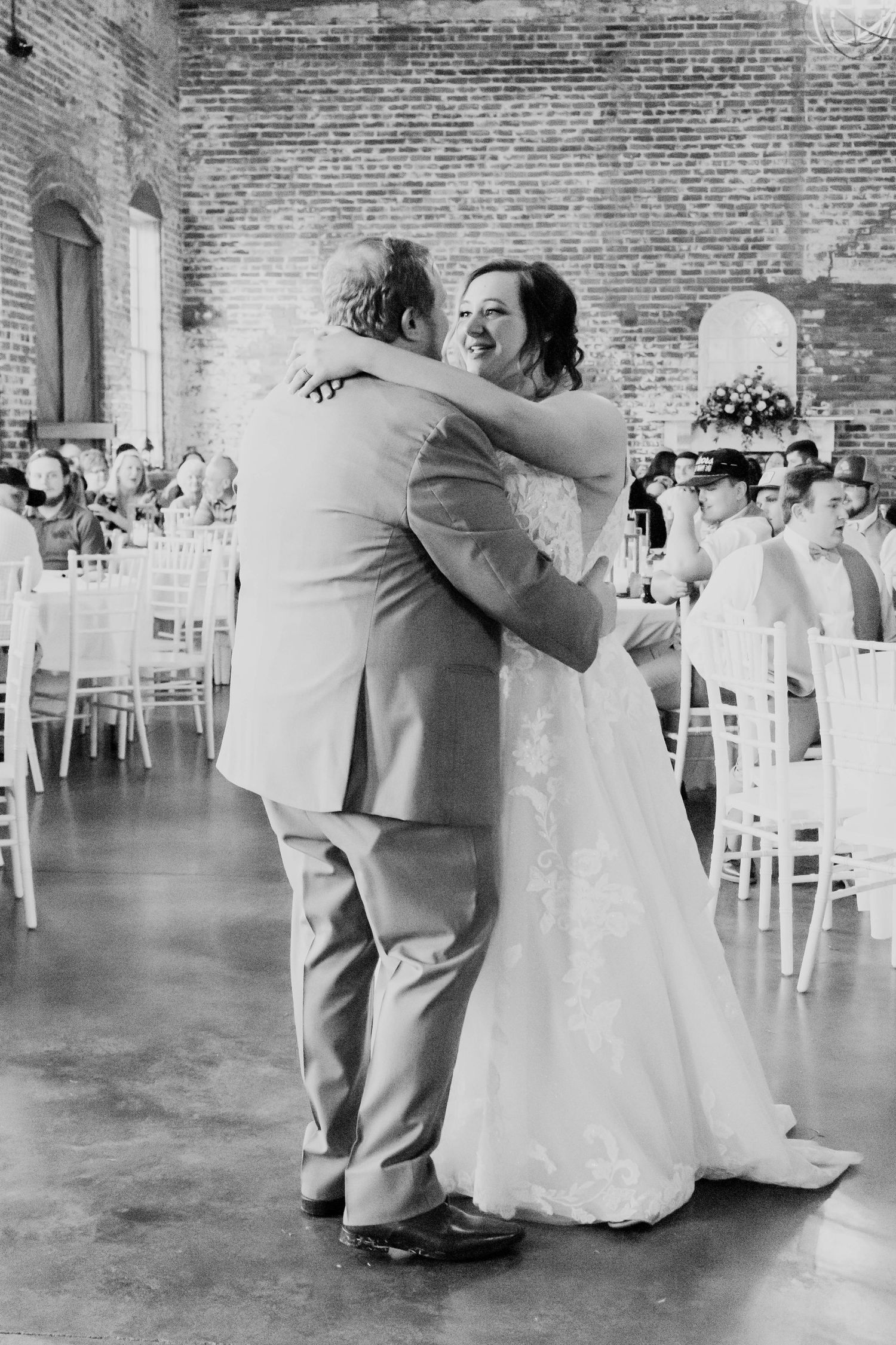Wedding couple dancing in a rustic brick-walled venue with white chairs in the background.