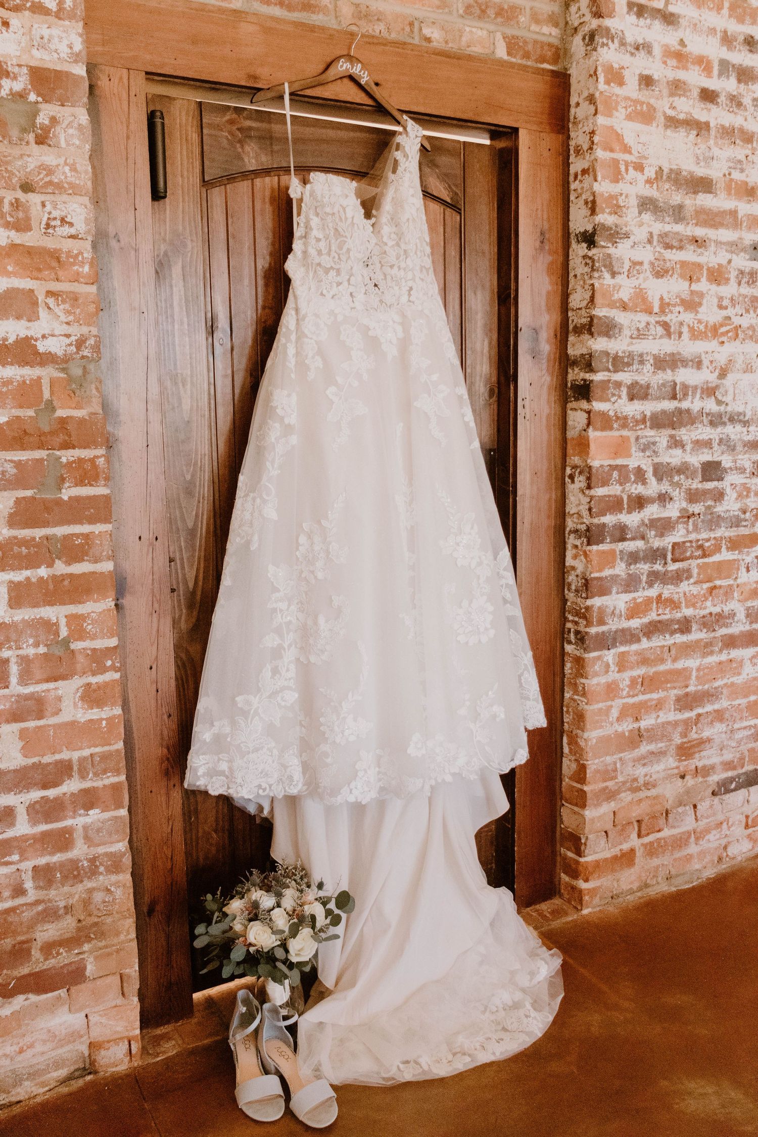 Elegant white lace wedding dress hanging on wooden door with bouquet and shoes at base.