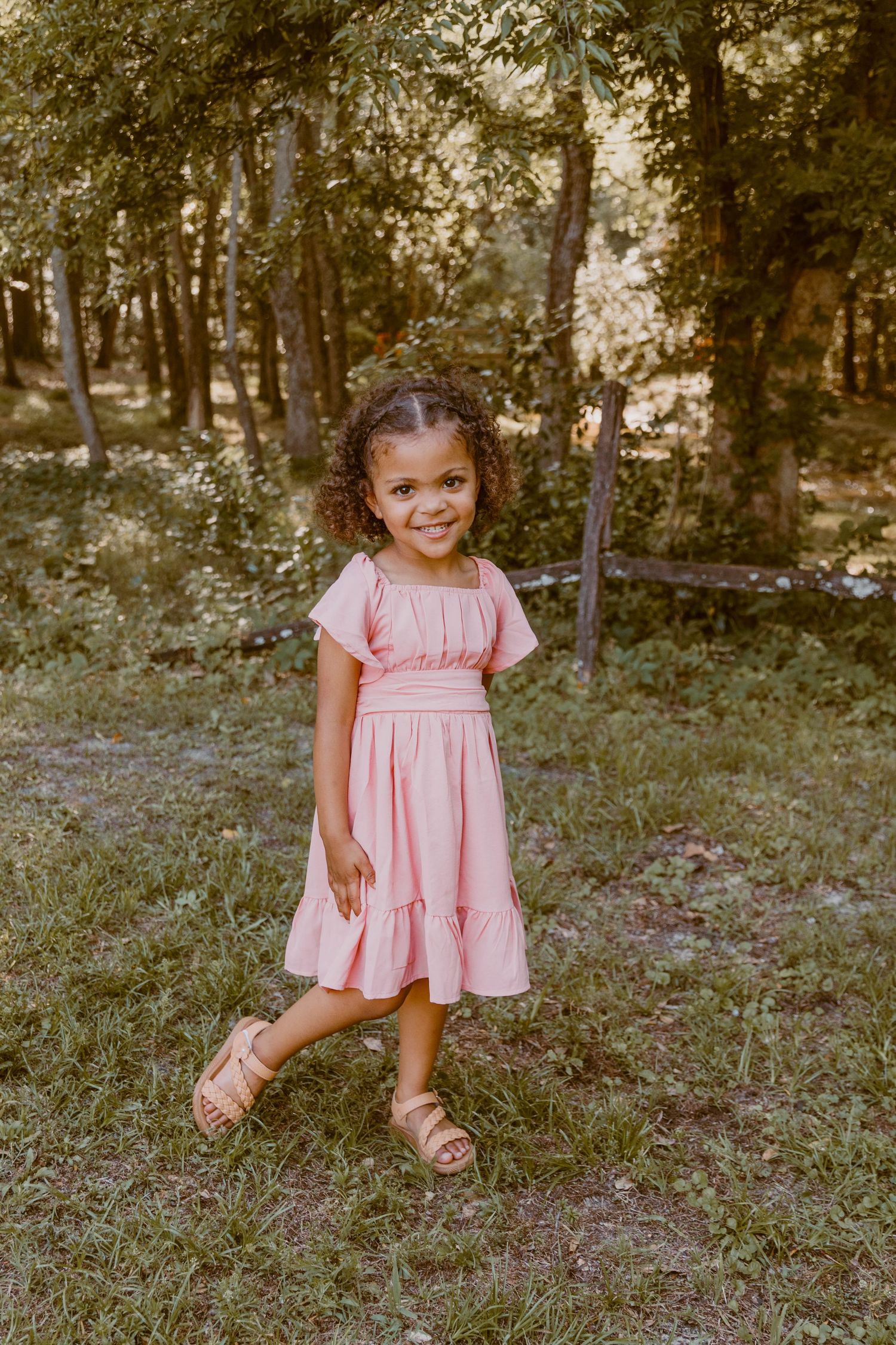 A child in a pink dress poses playfully in a wooded setting.