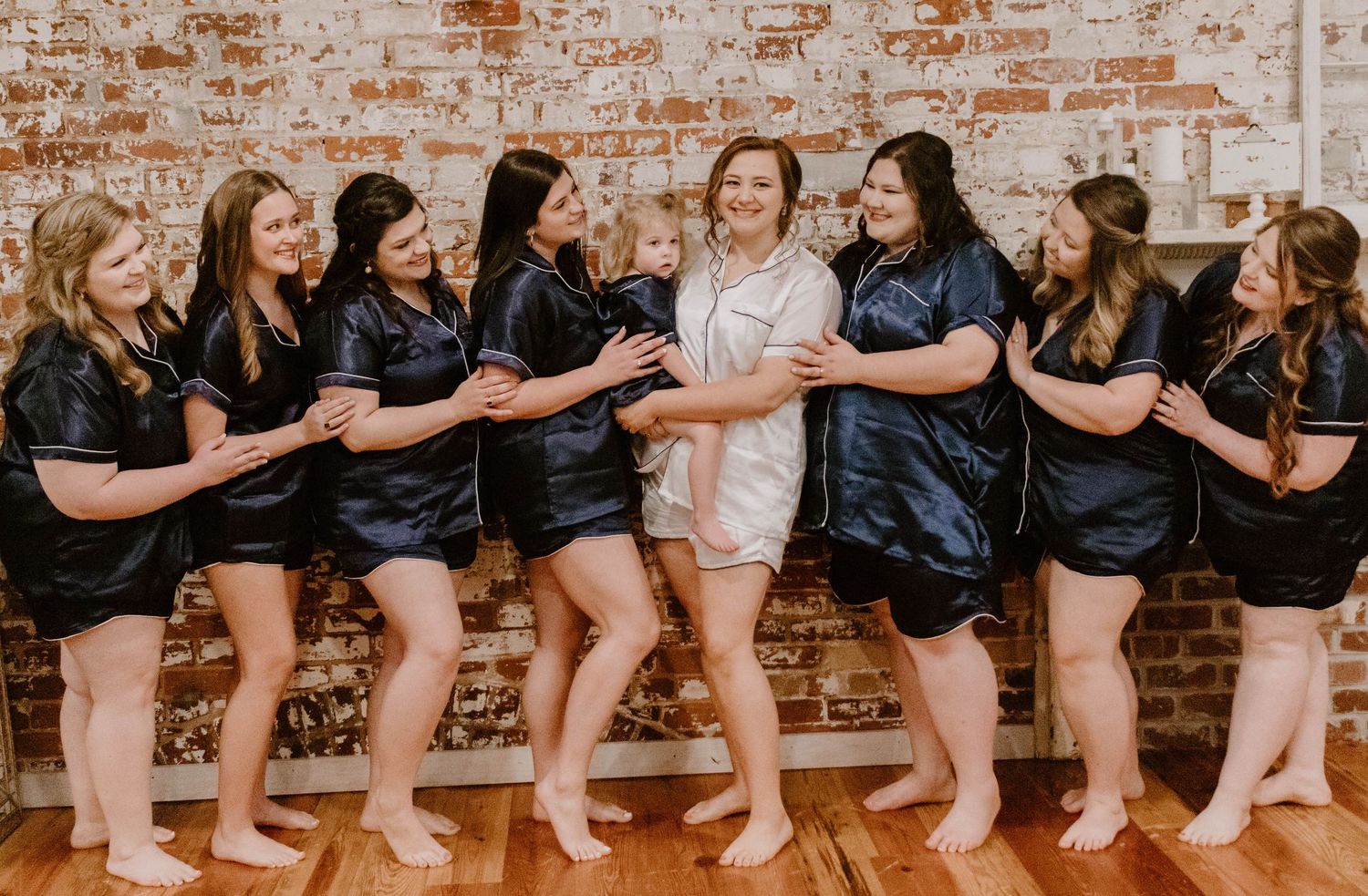 Row of bridesmaids sitting together showing their bare feet against brick wall.