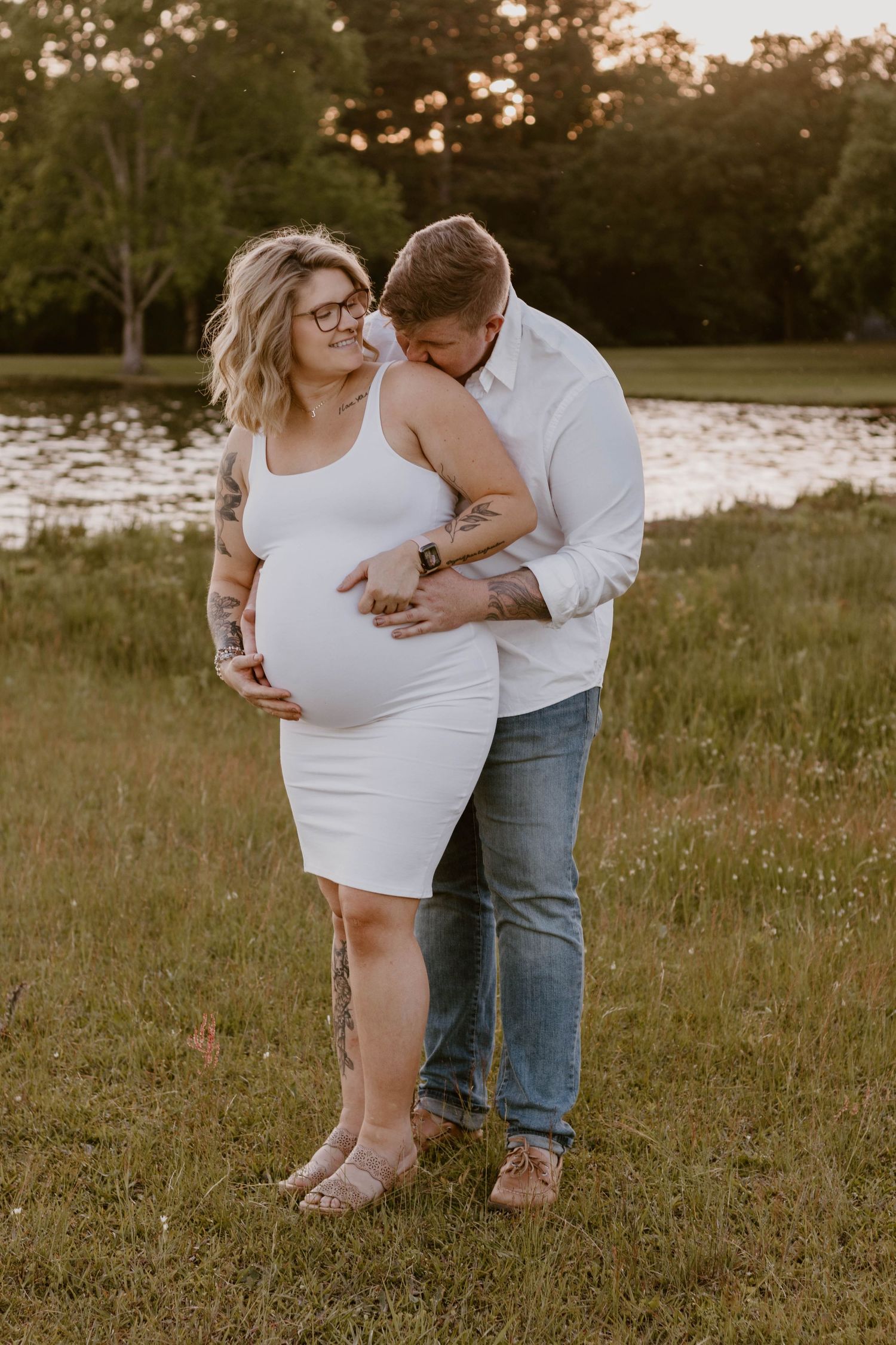 Maternity photo session in a grassy field at sunset showing couple embracing.