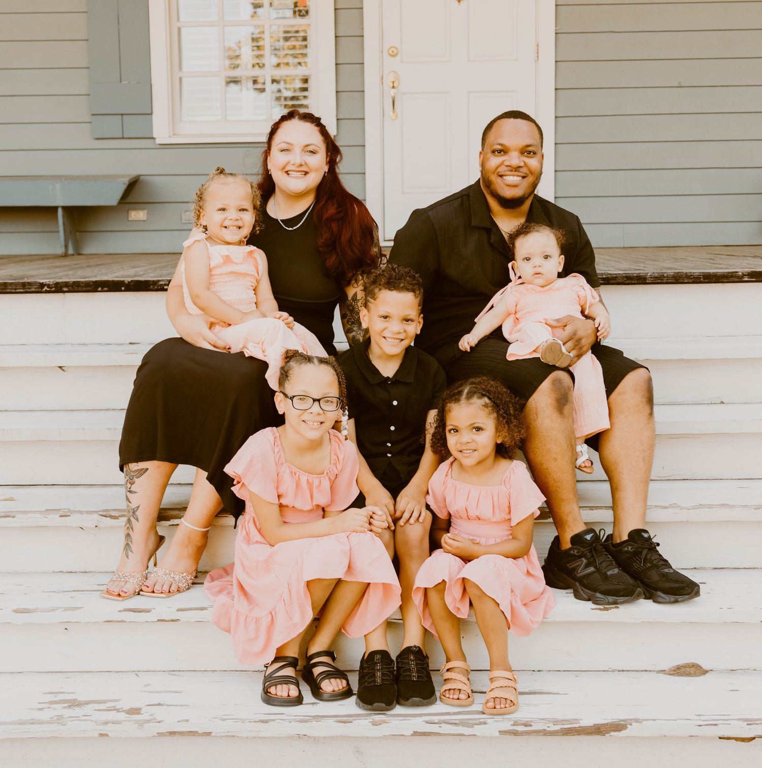 A family sits together on white porch steps wearing coordinated pink and black outfits.