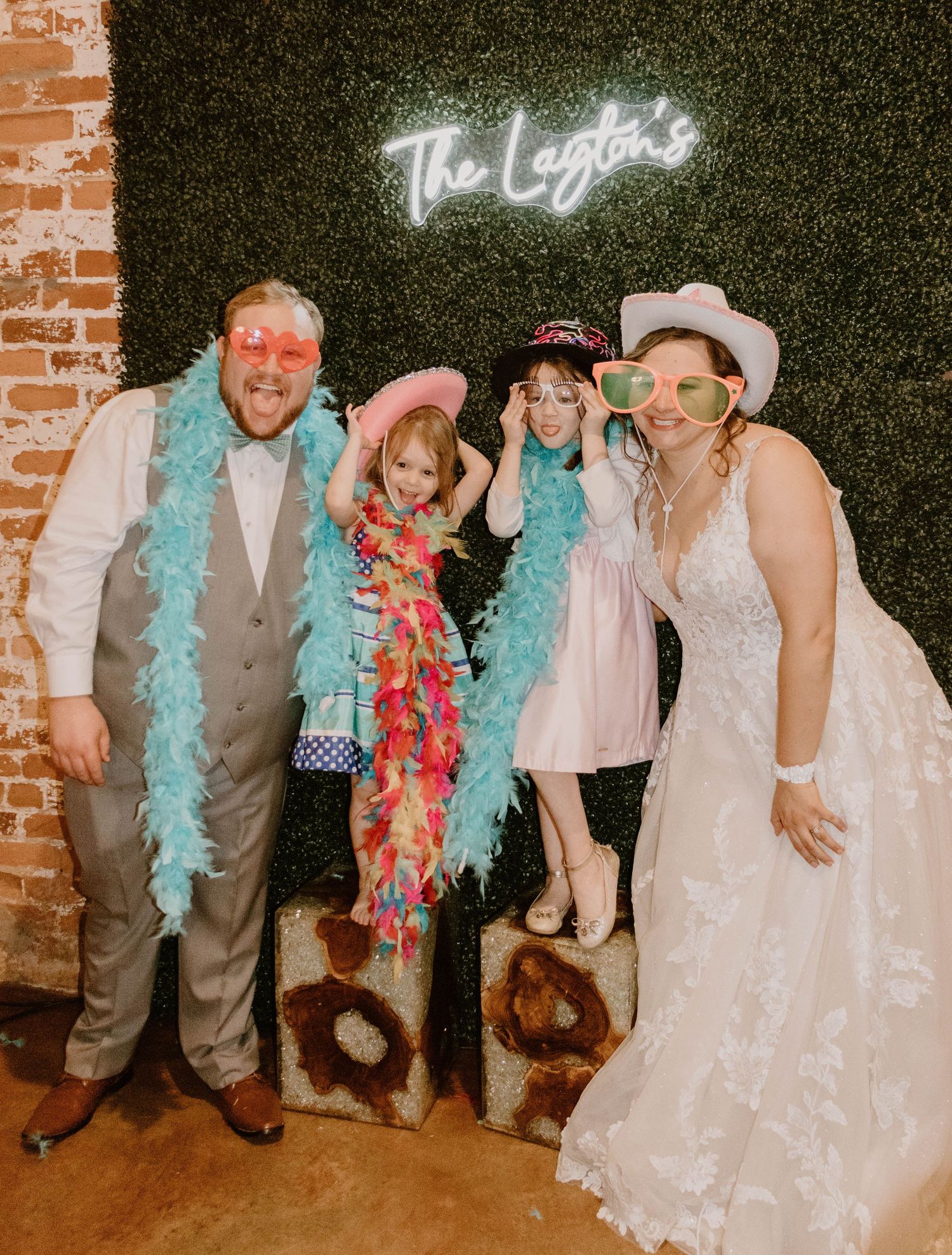 Wedding party members pose with colorful feather boas in front of a neon sign backdrop.