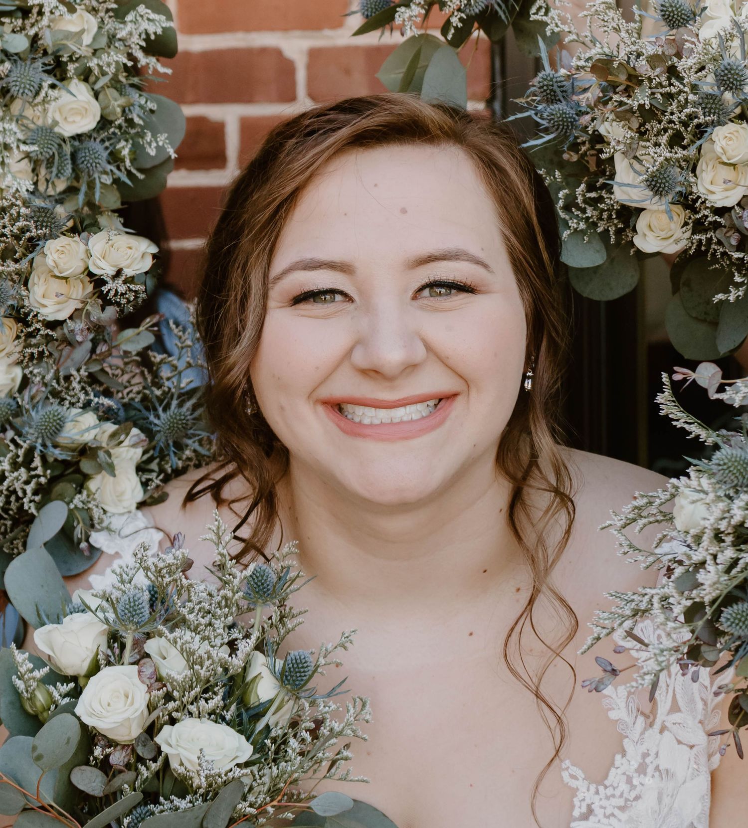 Wedding bouquet featuring white roses and dusty blue accents against brick background.