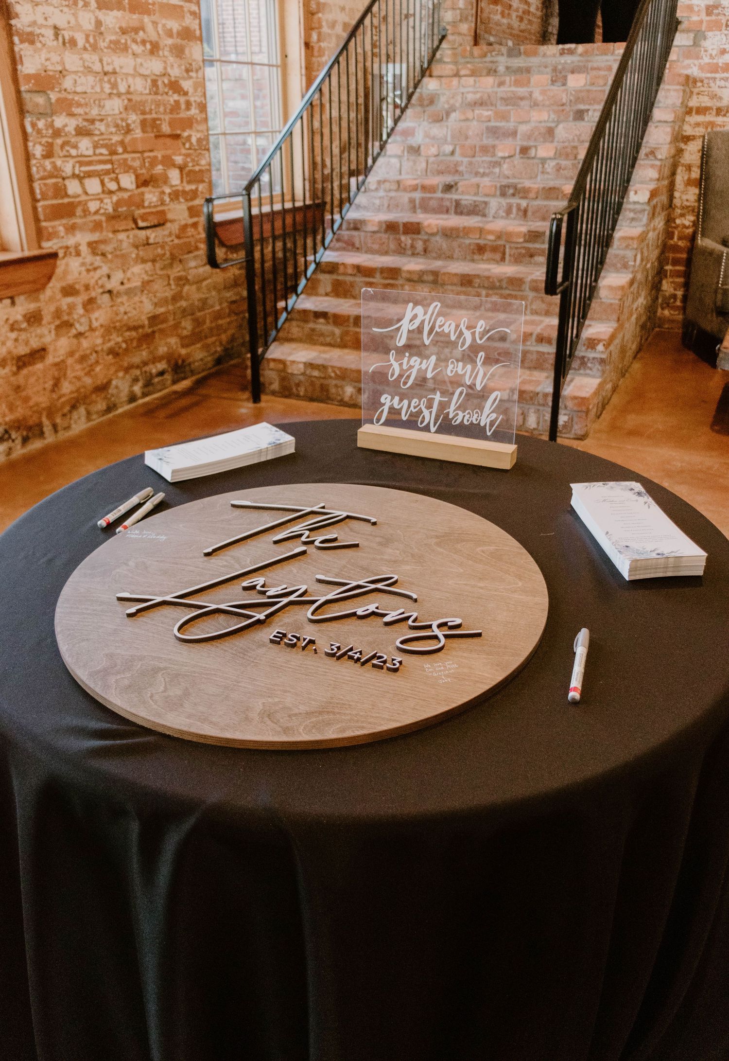 Wedding guest book signing station with wooden circular plaque on black tablecloth.
