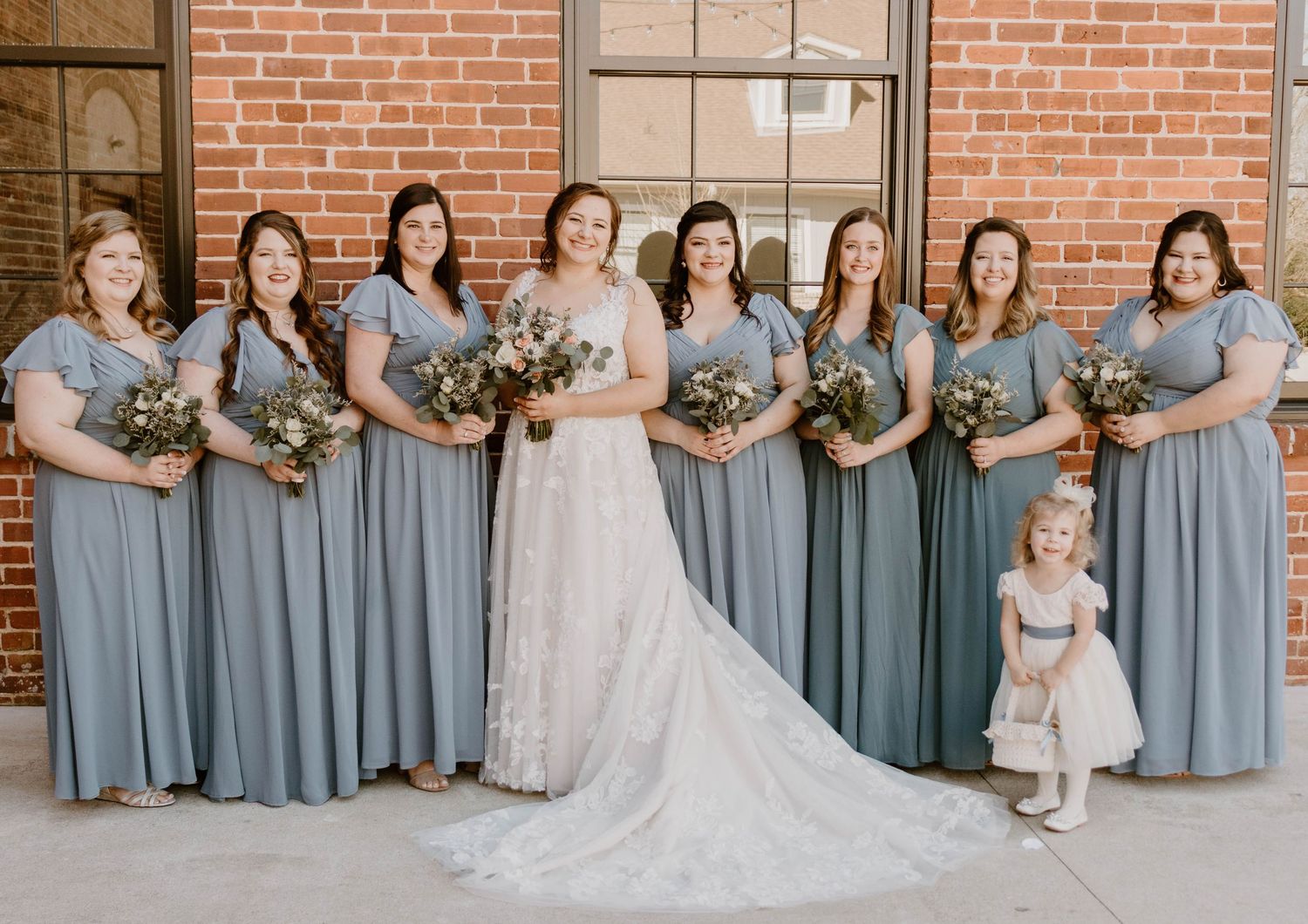 Wedding party in dusty blue bridesmaid dresses posing against brick wall.
