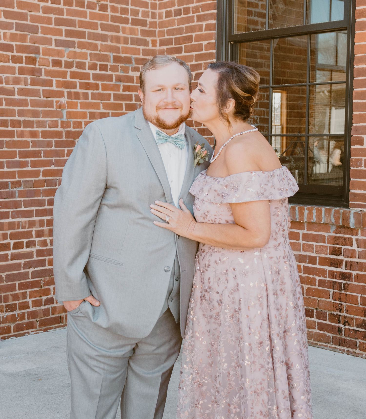 Couple in gray suit and pink dress share intimate moments against brick wall backdrop.