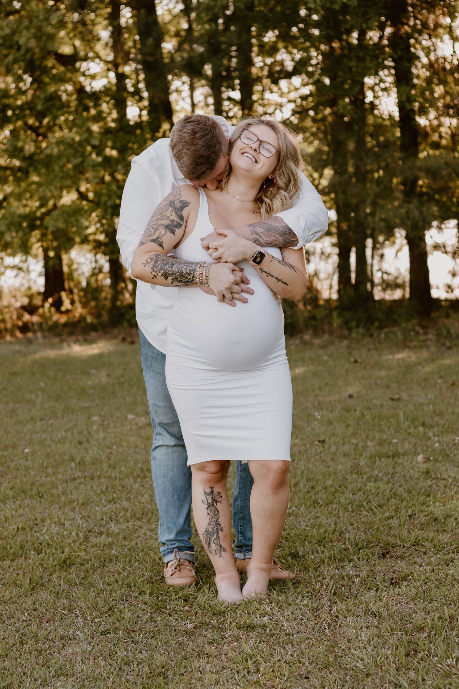 A loving embrace is captured during a maternity photoshoot in a wooded setting at sunset.