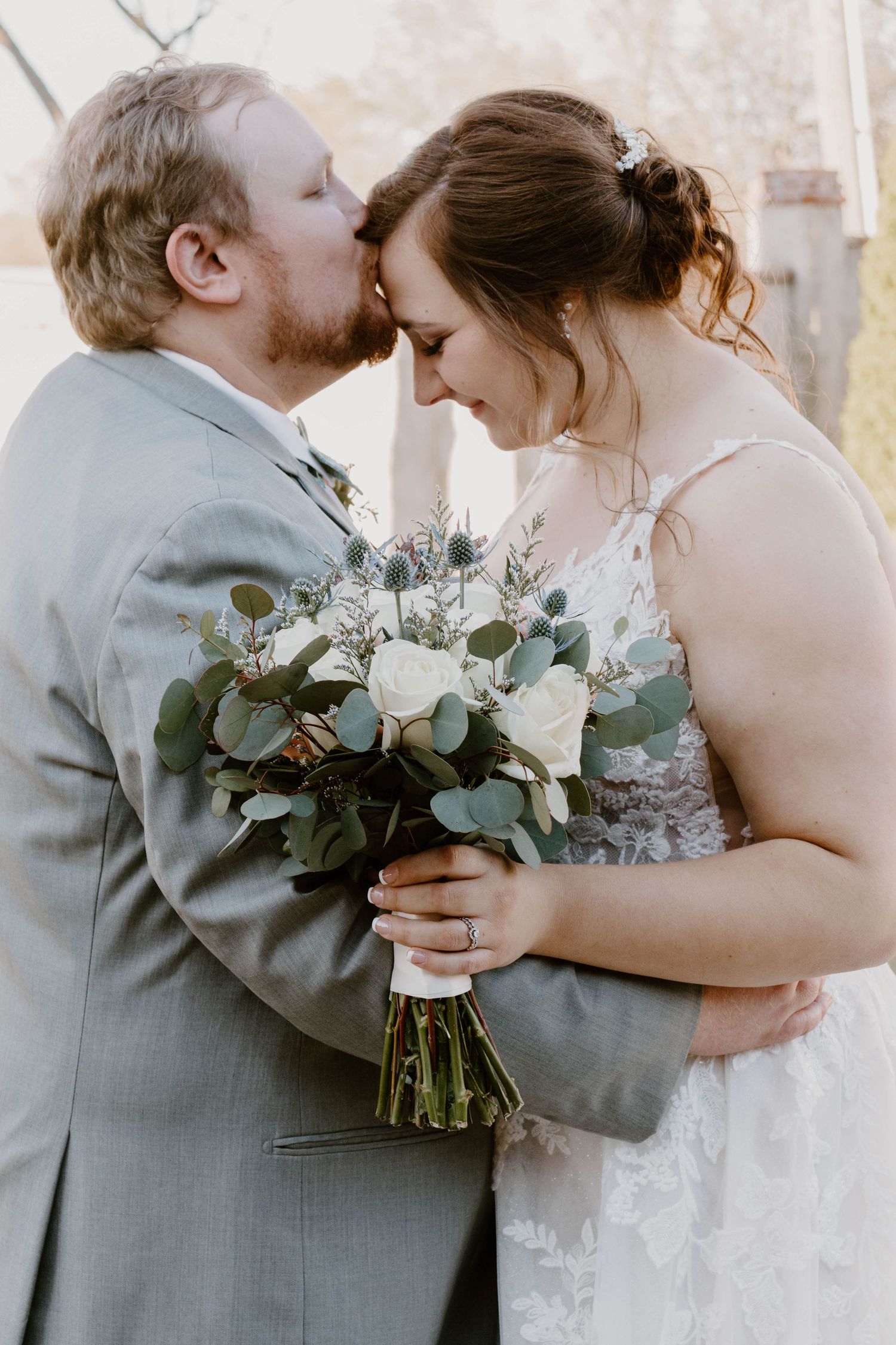Romantic wedding couple embraces while holding a white and green floral bouquet against a textured wall.