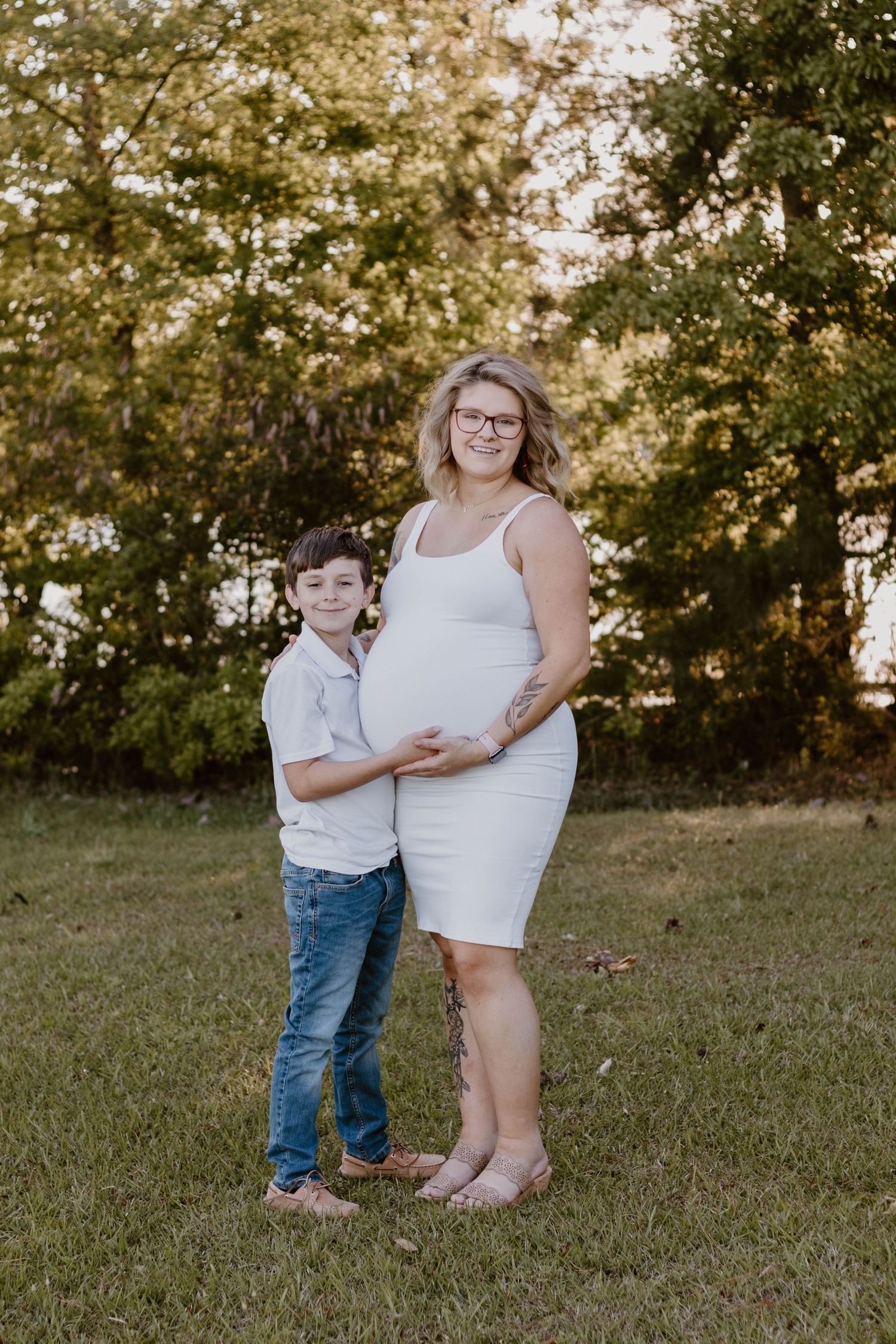 A young boy hugs his mother's pregnant belly while standing in an outdoor setting.
