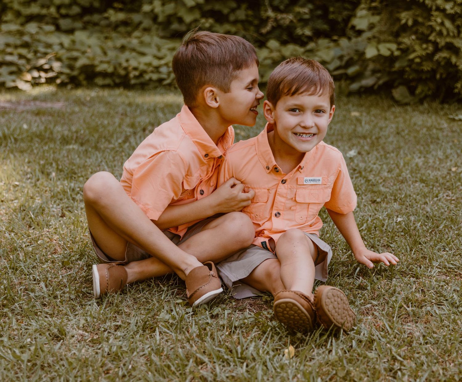 Two children in peach shirts playing and laughing while sitting on grass.
