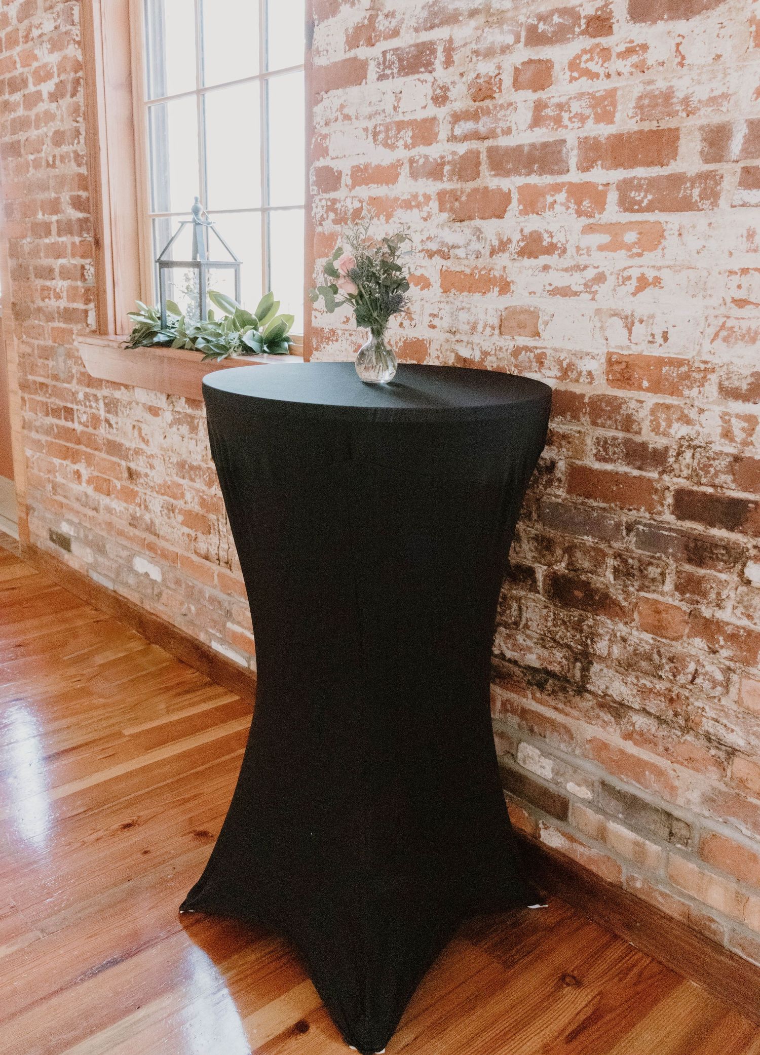 A tall black cocktail table with spandex cover against exposed brick wall with decorative plants on windowsill.