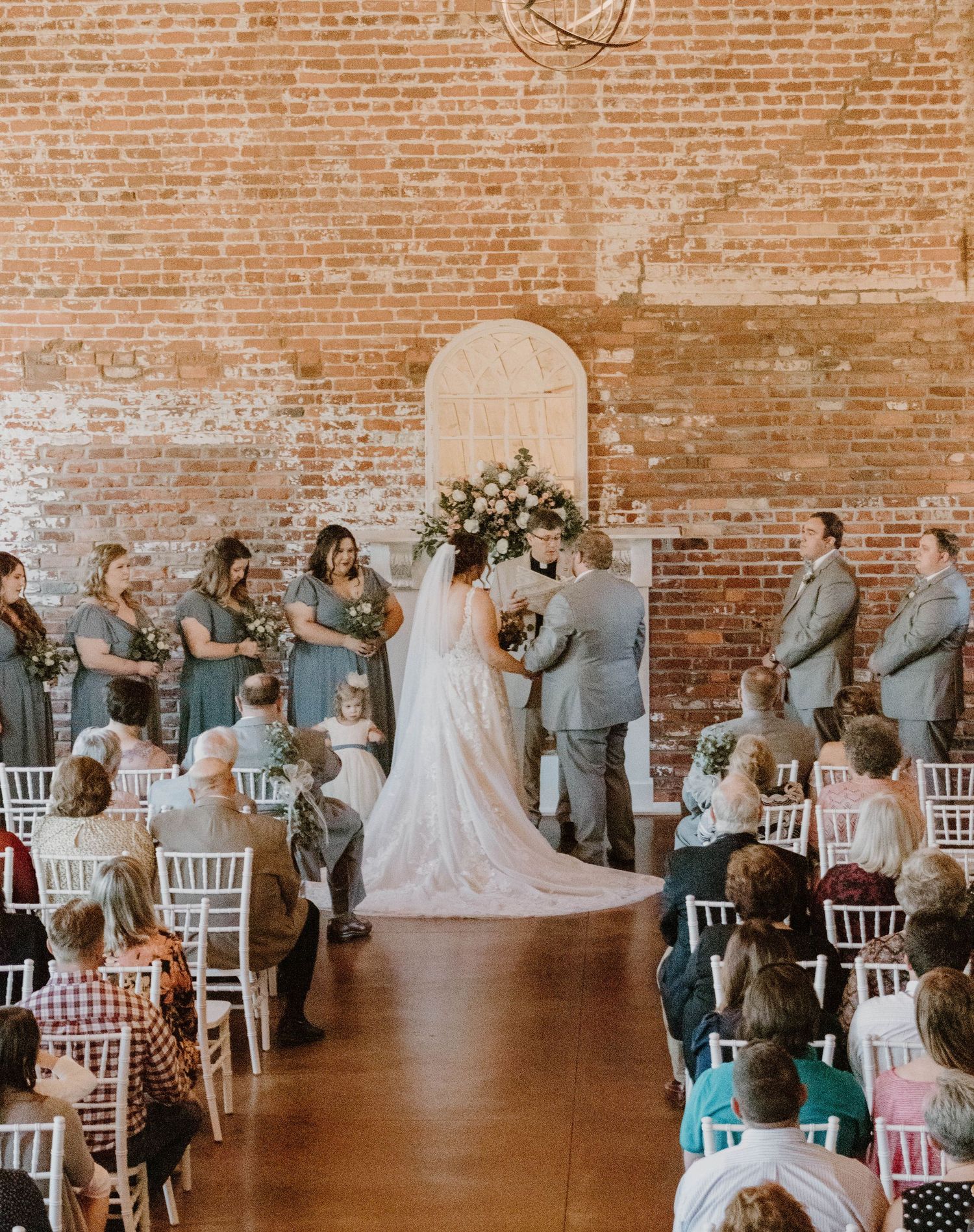 Wedding party celebrates in an industrial brick venue with white chairs and romantic decor.