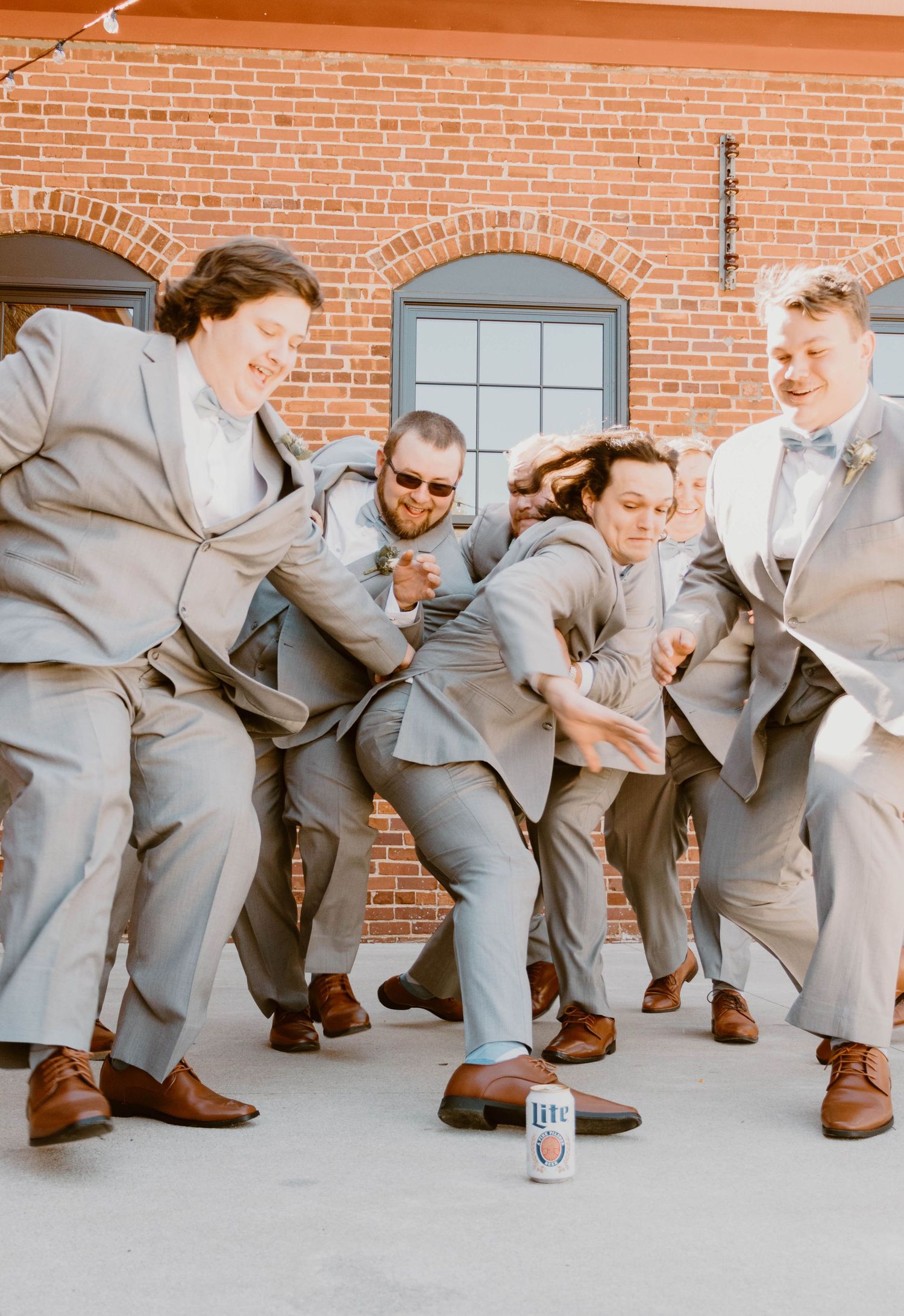 Groomsmen in gray suits perform a playful bowling pose sequence against a brick wall.