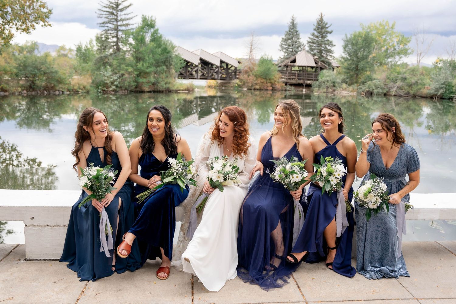 Wedding party in navy blue dresses sits by a scenic lake sharing candid moments together during golden hour.