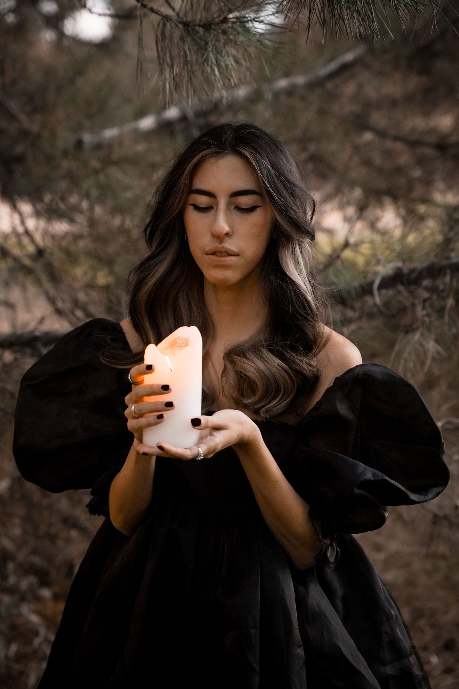 Atmospheric portrait with glowing candle in dark forest setting wearing black clothing.
