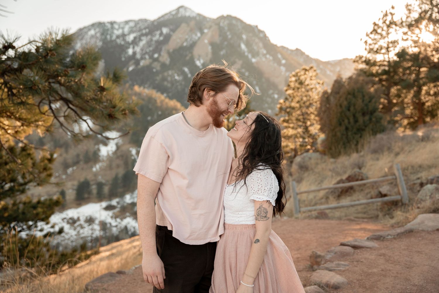 Couple sharing a romantic kiss outdoors with mountain and pine tree backdrop in golden hour light.