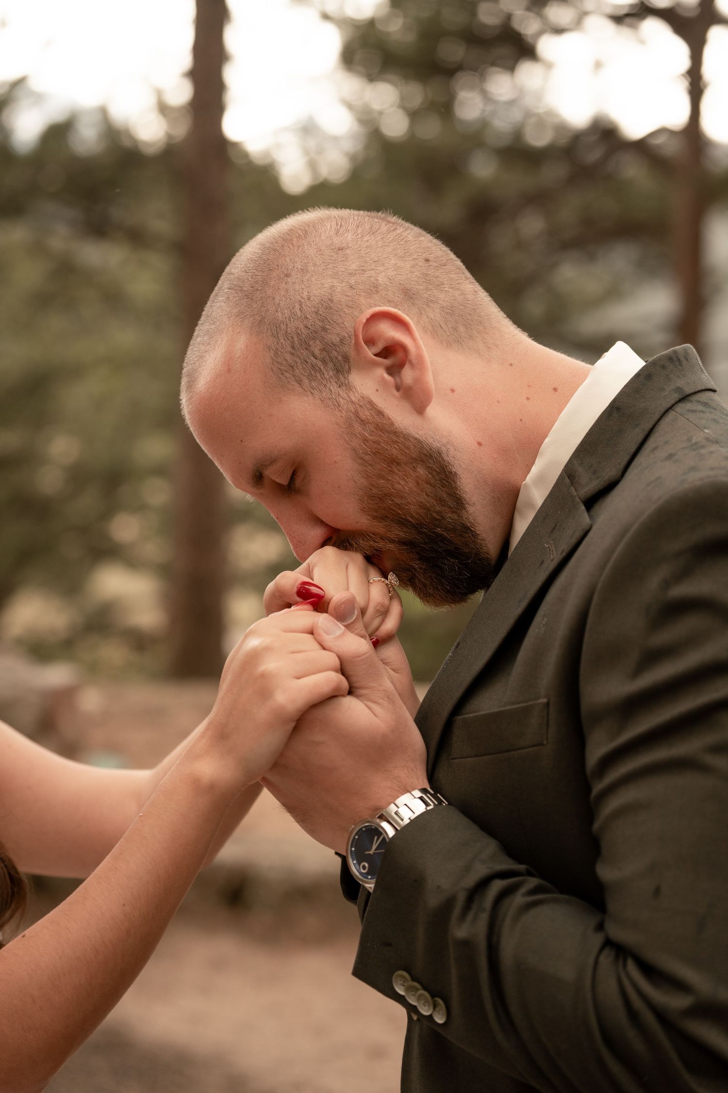 Bearded groom in dark suit tenderly kisses bride's hand during outdoor wedding ceremony in pine forest.