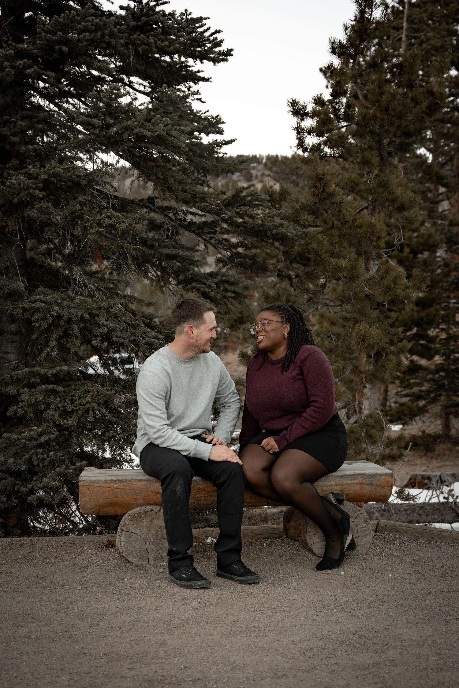 A couple sits together on a wooden bench surrounded by snow-covered pine trees.