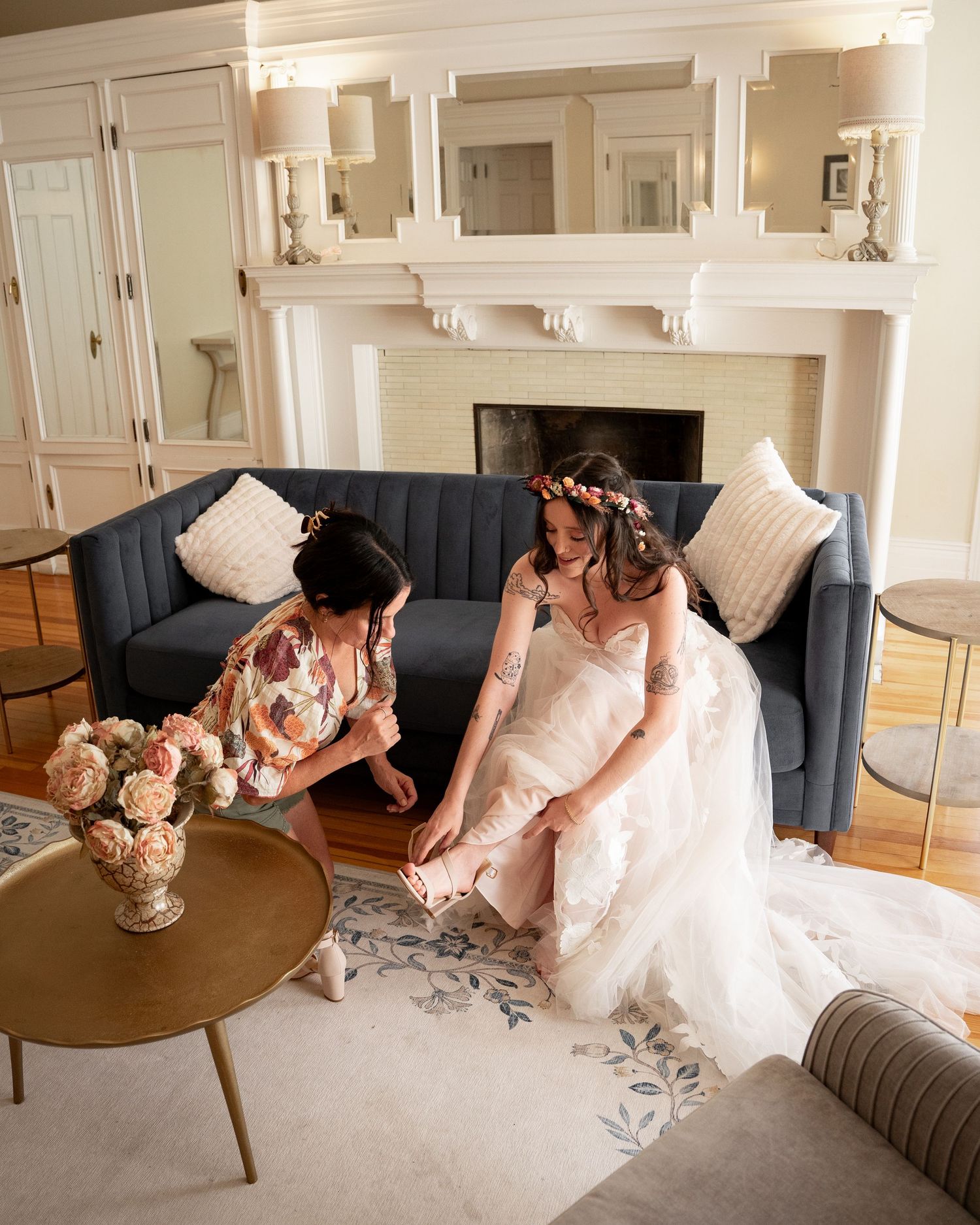 Bride to be wearing a pink flower crown is assisted by her mother in putting on her shoes as she gets ready for her wedding day.