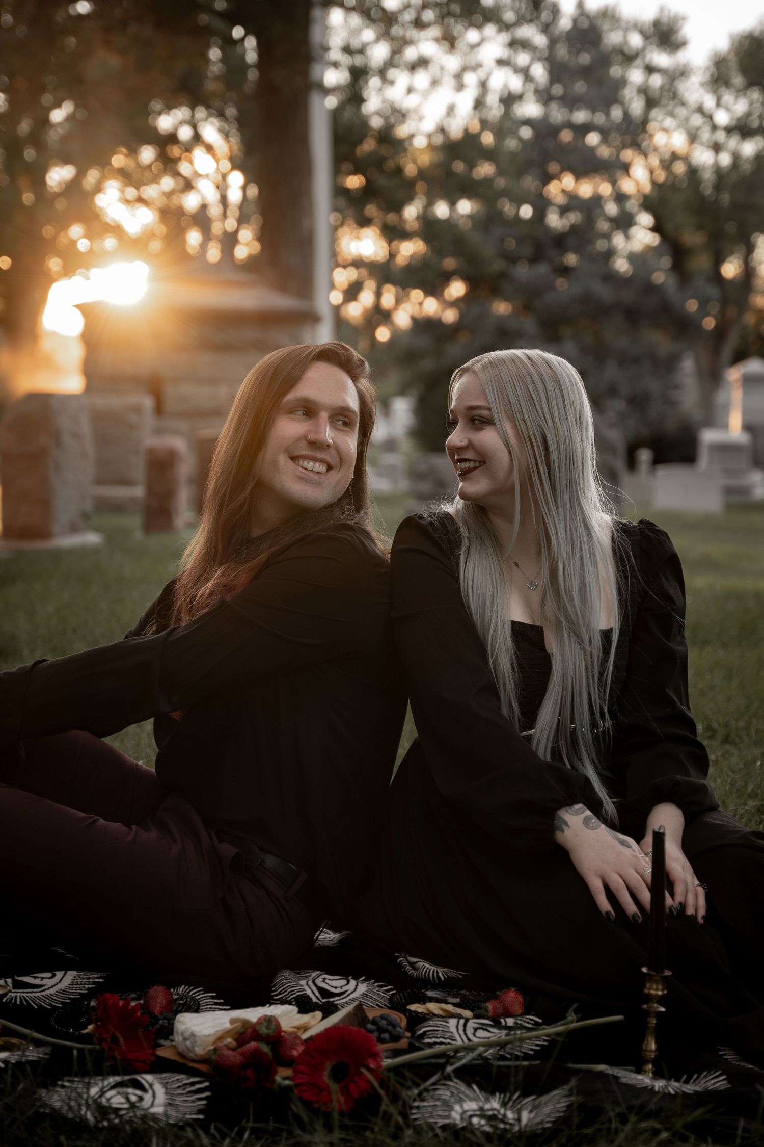 A couple dressed in black sit together on a blanket in a cemetery at golden hour sunset.