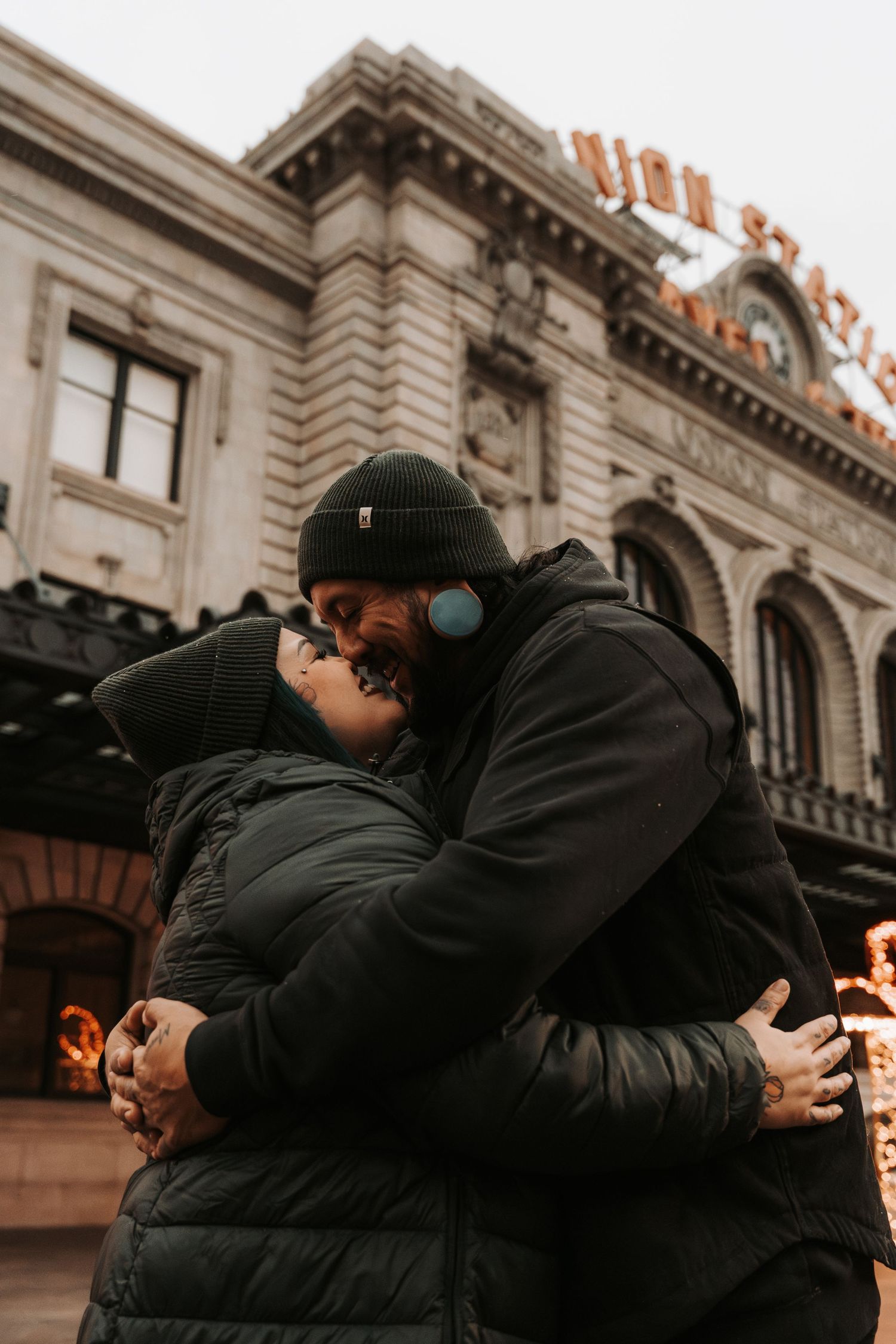 Couple embracing and kissing in front of Union Station building, wearing winter jackets and beanies on a cloudy day.