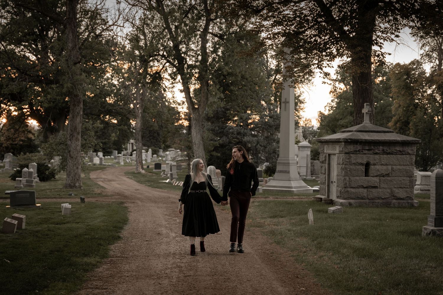 Two figures in vintage attire walk along a path in a historic cemetery at dusk.