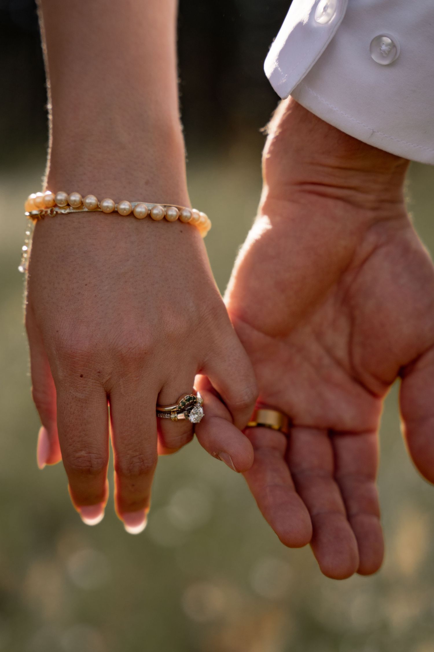 Two hands wearing wedding rings and pearl bracelets reach out to hold each other in warm sunlight.