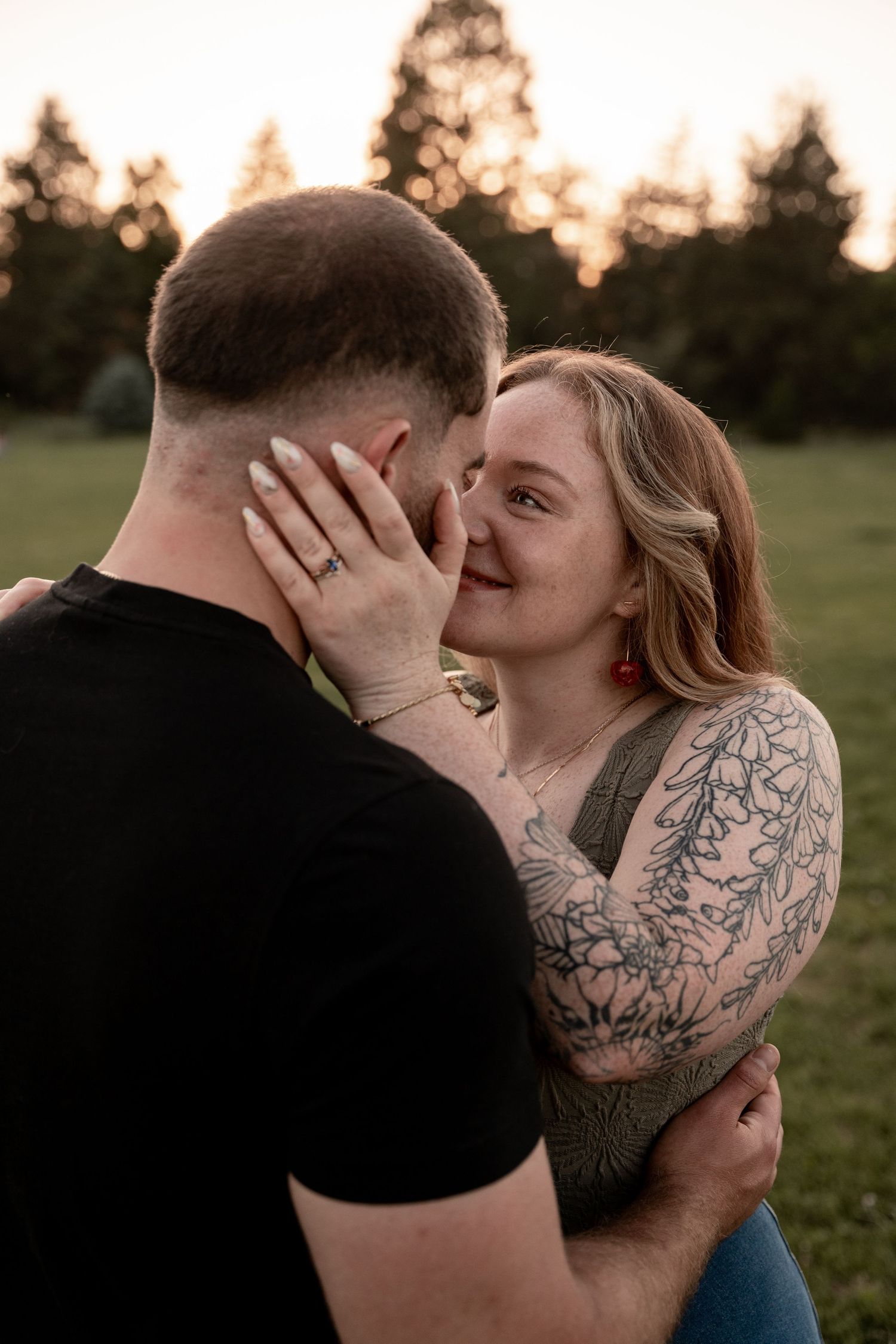 A tattooed woman smiles while embracing a man in a black shirt outdoors at golden hour.