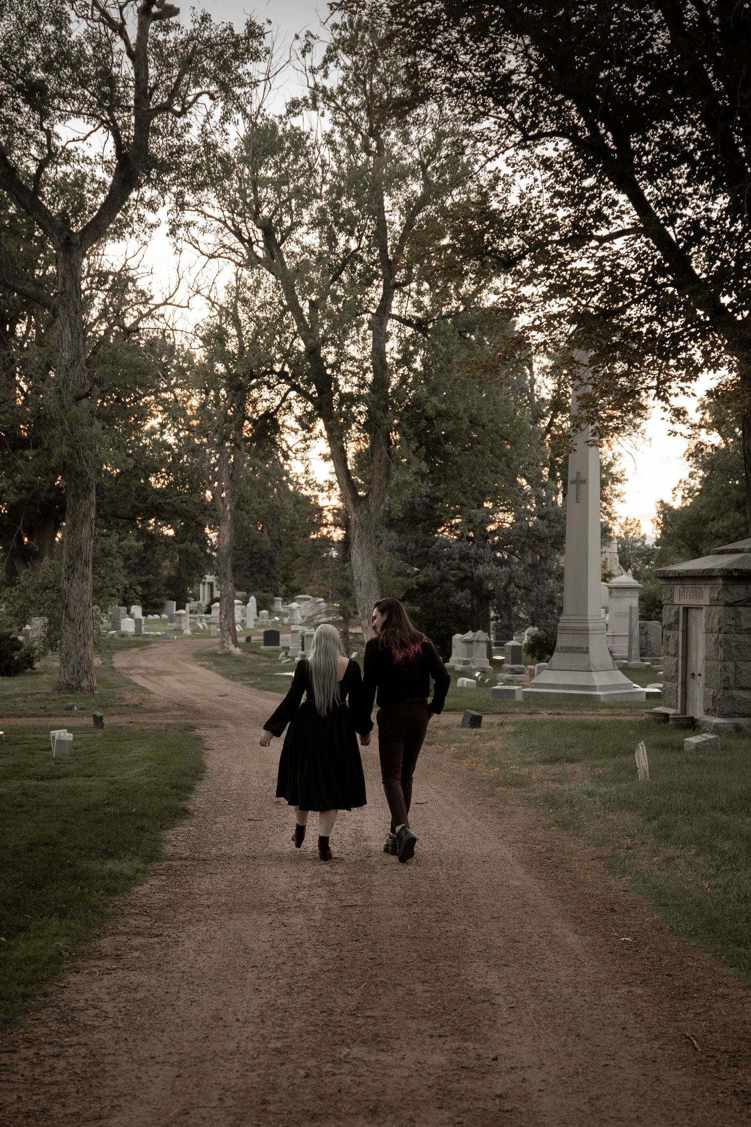 Two figures in dark clothing walk along a cemetery path at dusk, surrounded by tombstones and tall trees.