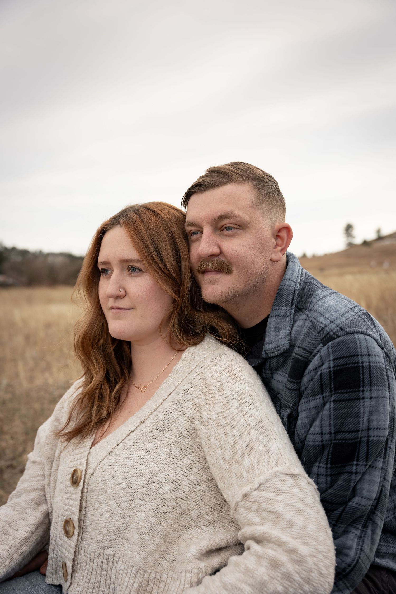 A couple sits together in a golden field, the woman in a cream sweater leaning back against a man in a plaid shirt.