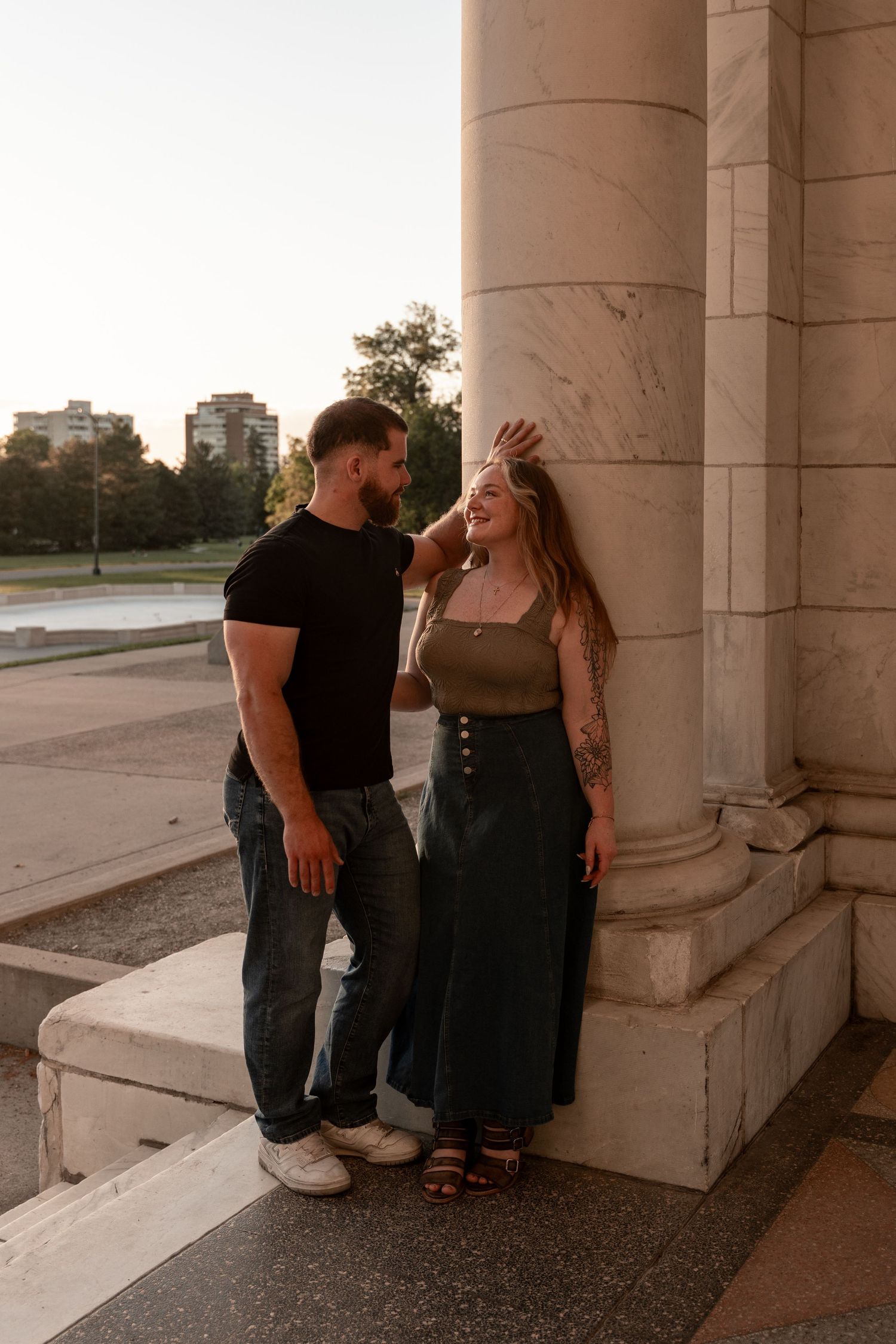 A couple shares a romantic moment leaning against a marble column outdoors at sunset, woman with tattoos laughing joyfully.