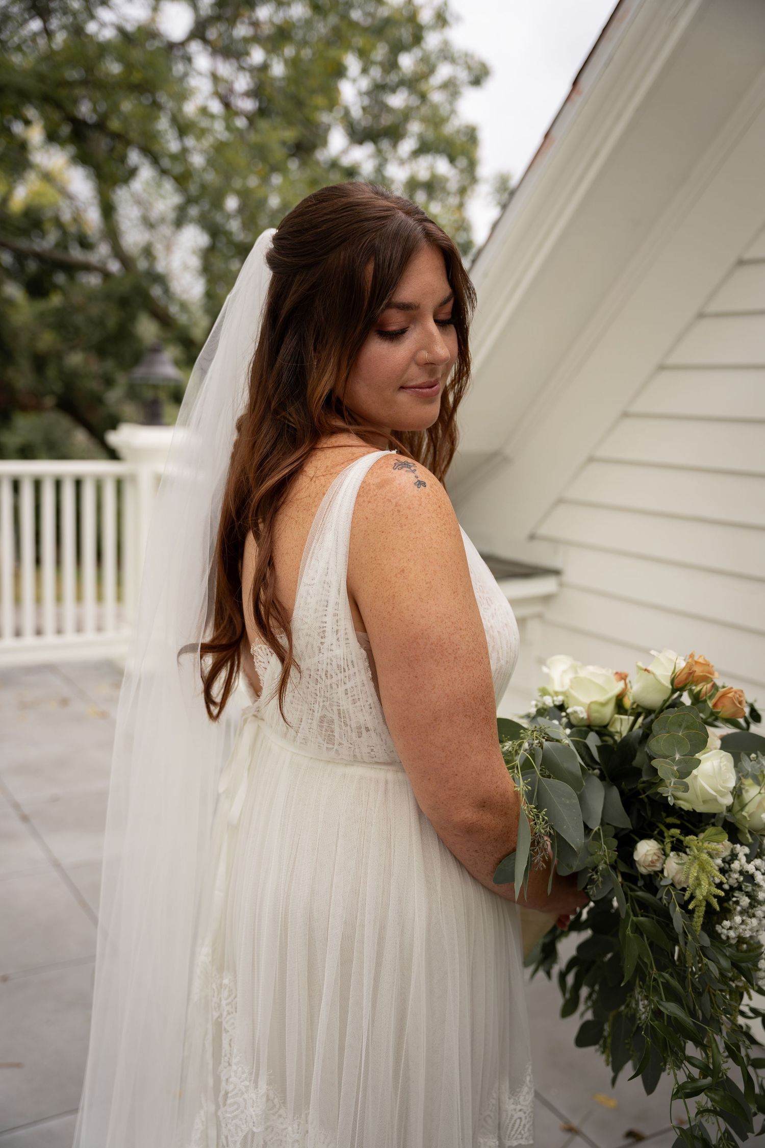 A bride in a white lace wedding dress holds a lush bouquet of white flowers and greenery on a porch.