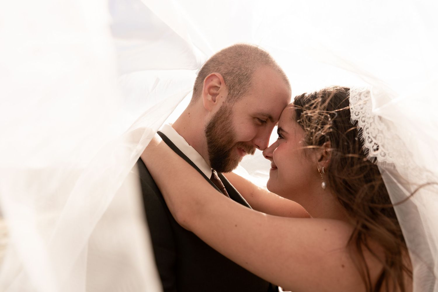 A bride and groom share an intimate forehead-to-forehead moment under a flowing white veil on their wedding day.