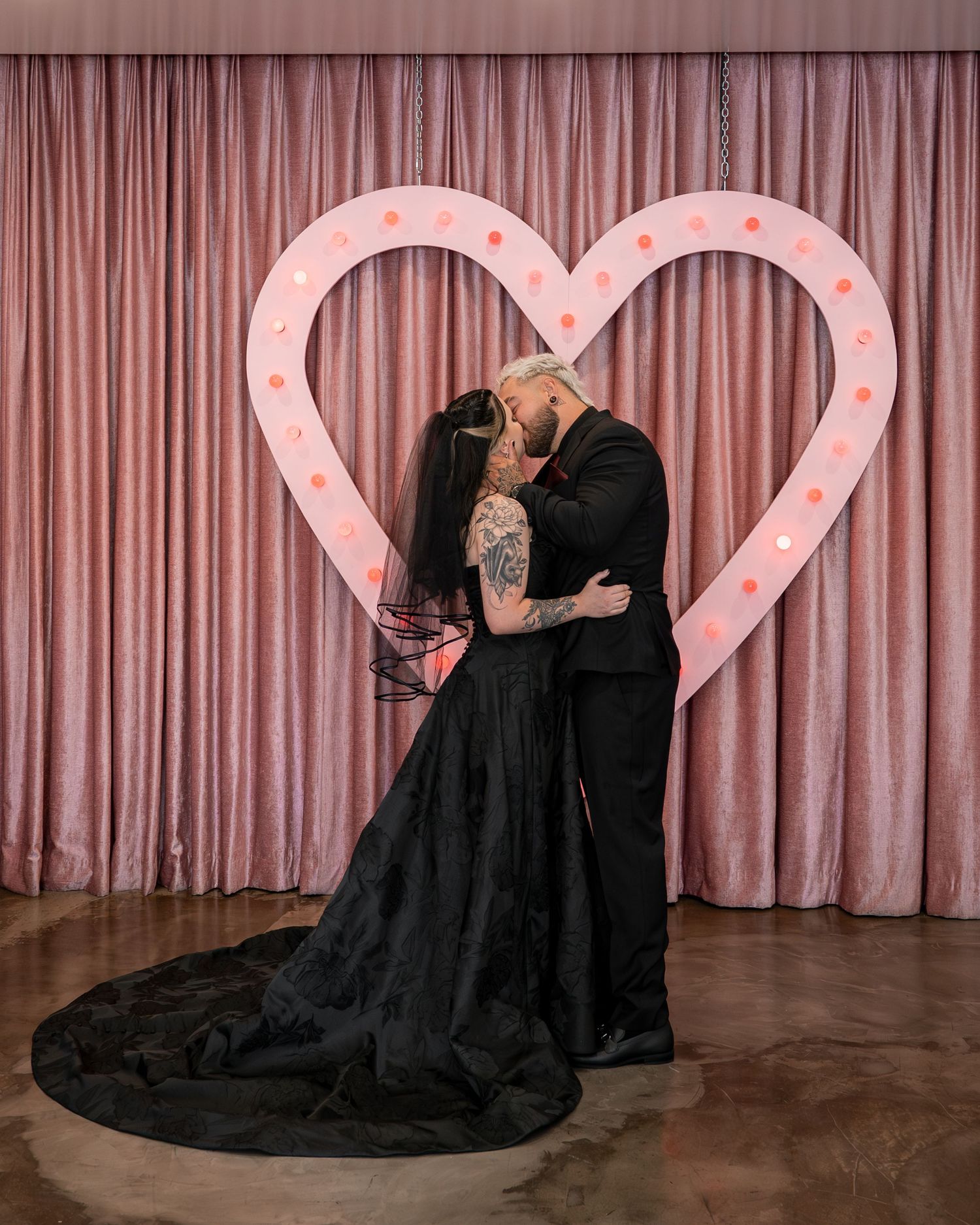 Couple shares a dramatic kiss against a pink curtain backdrop with illuminated heart decorations.