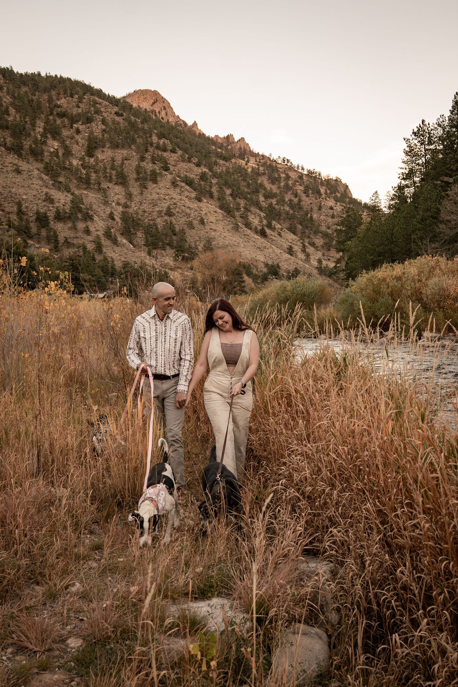Couple walking through tall grass with mountains and autumn foliage in the background.