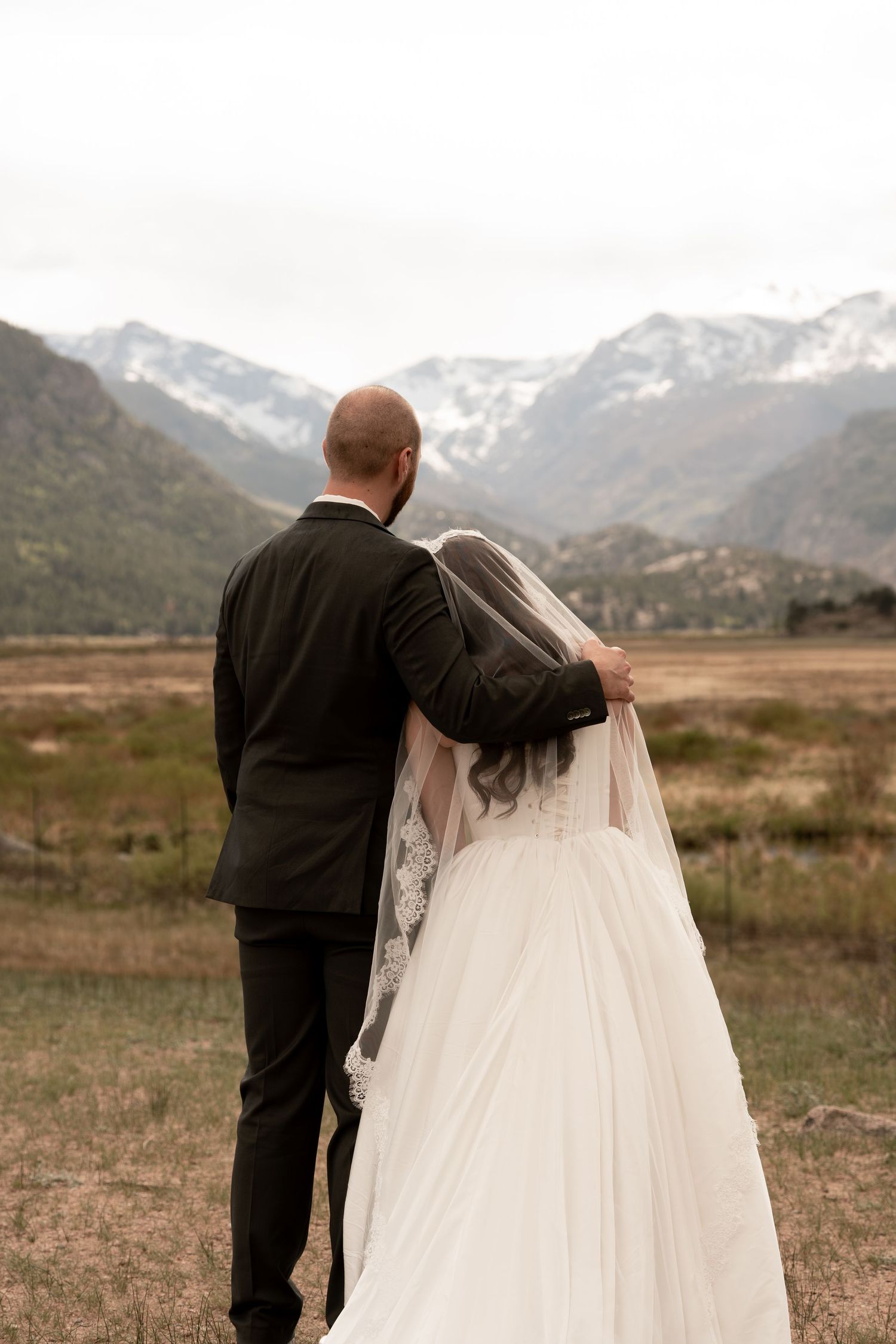 Bride and groom viewed from behind admiring snow-capped Rocky Mountains in wedding attire.