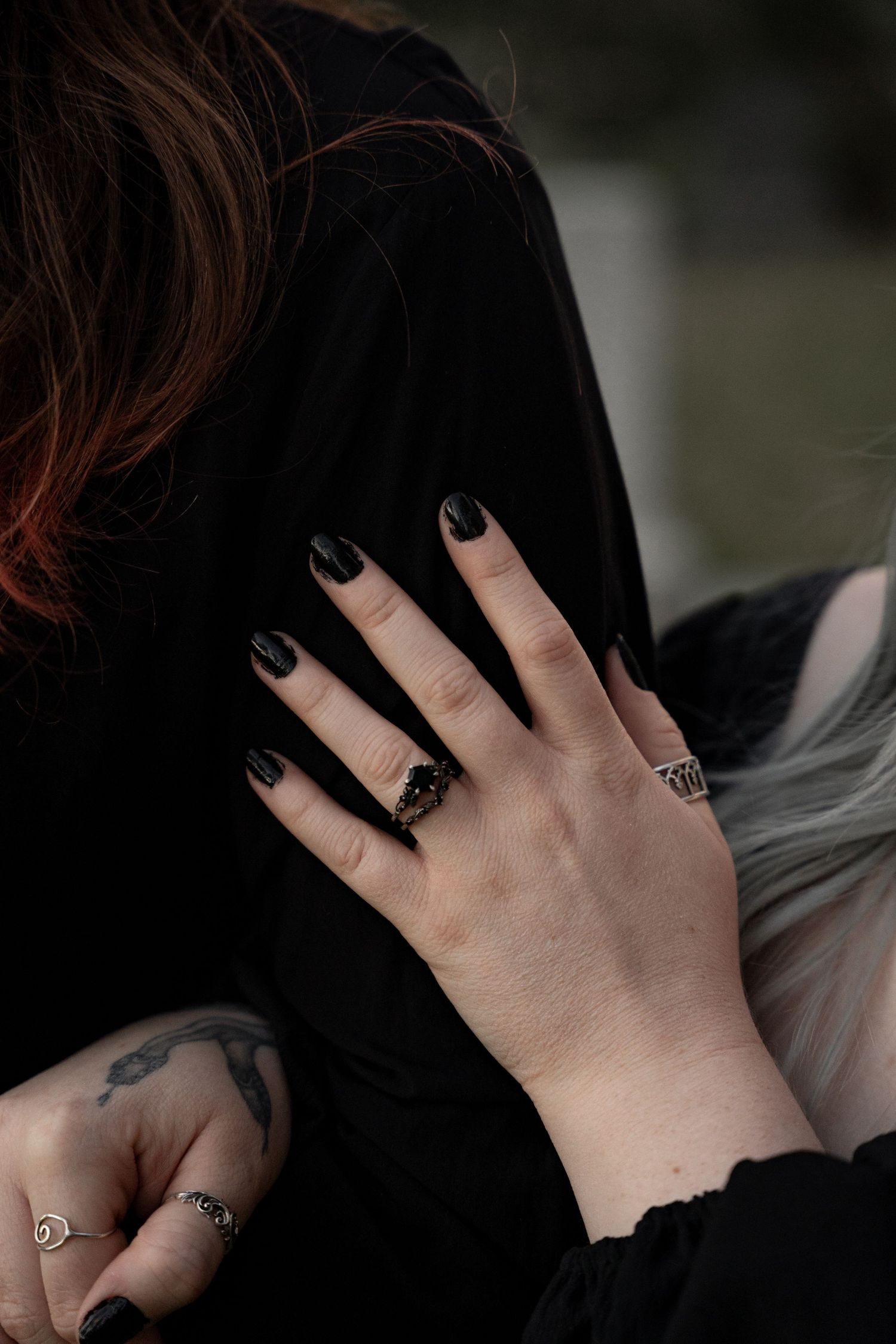 Hand with black nails and dark rings resting on a person wearing black, with a tattoo visible on the wrist.