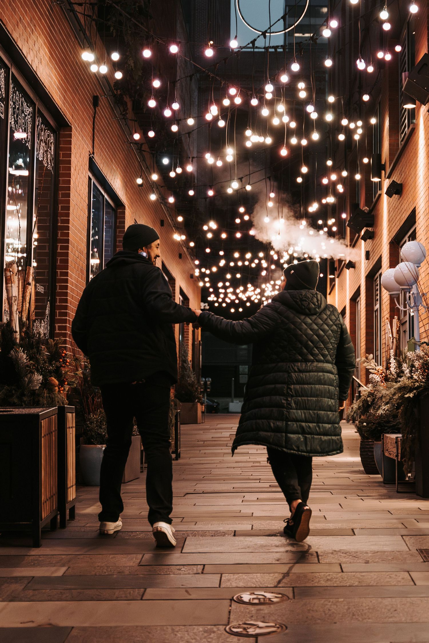 Couple walking hand-in-hand through a cozy alley adorned with warm string lights on a cold winter night.