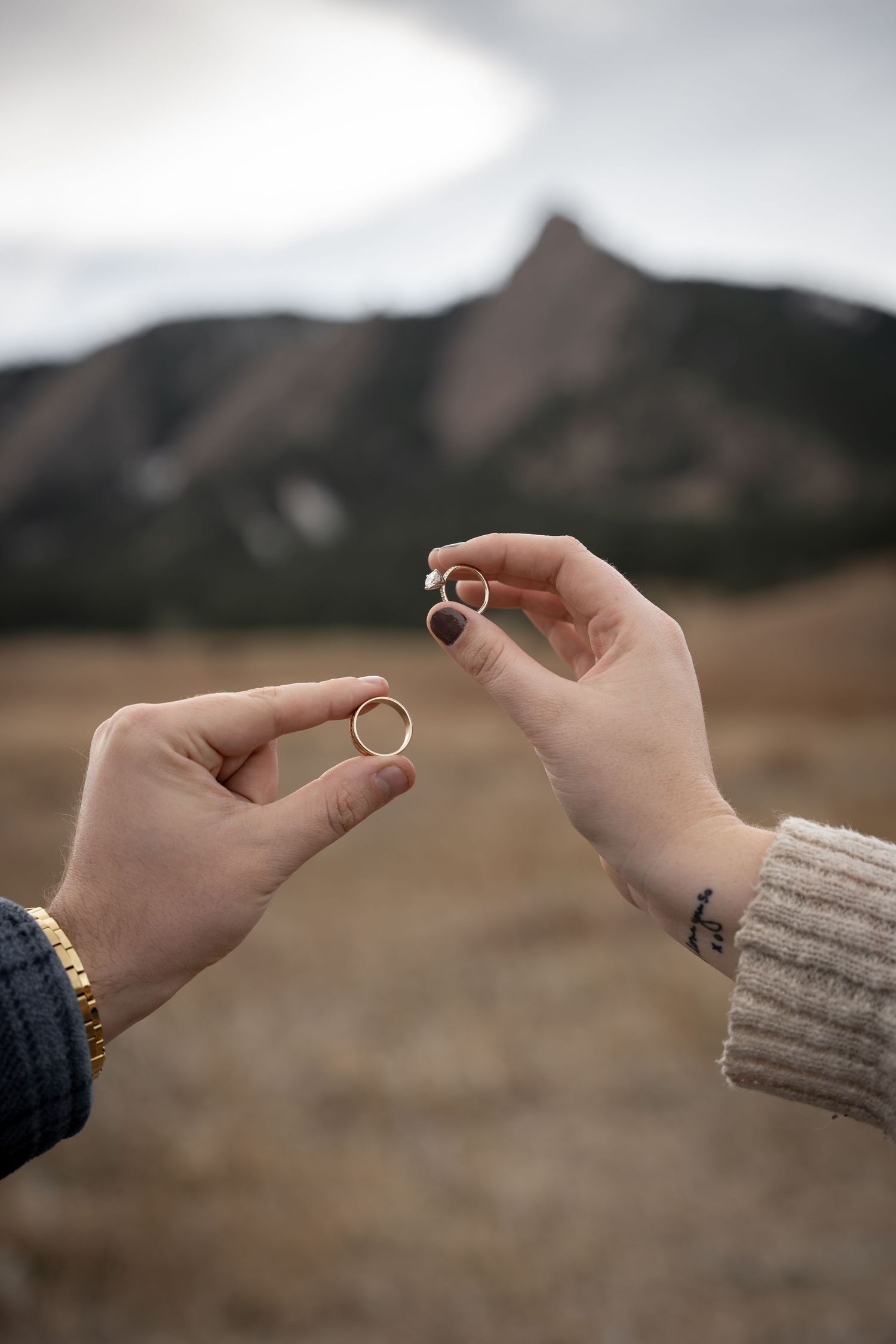 Two hands holding gold wedding rings outdoors with rocky mountain peaks in the background.