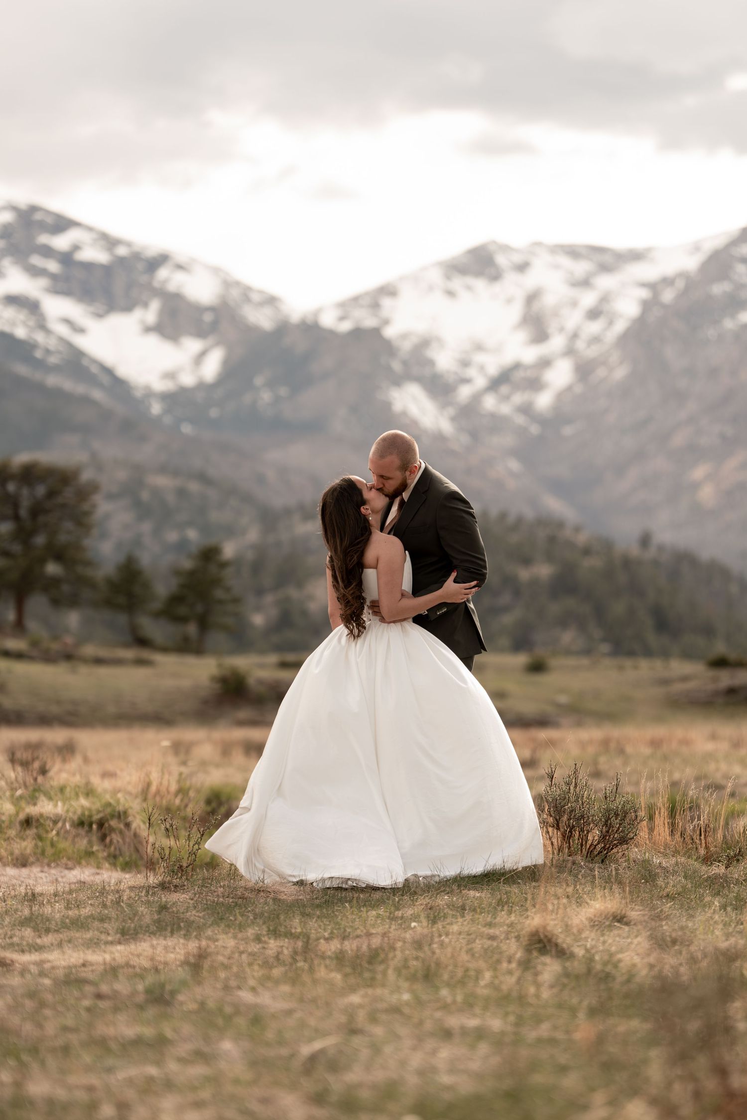 Bride and groom share a romantic kiss in a mountain meadow, with snow-capped peaks and cloudy skies behind them.