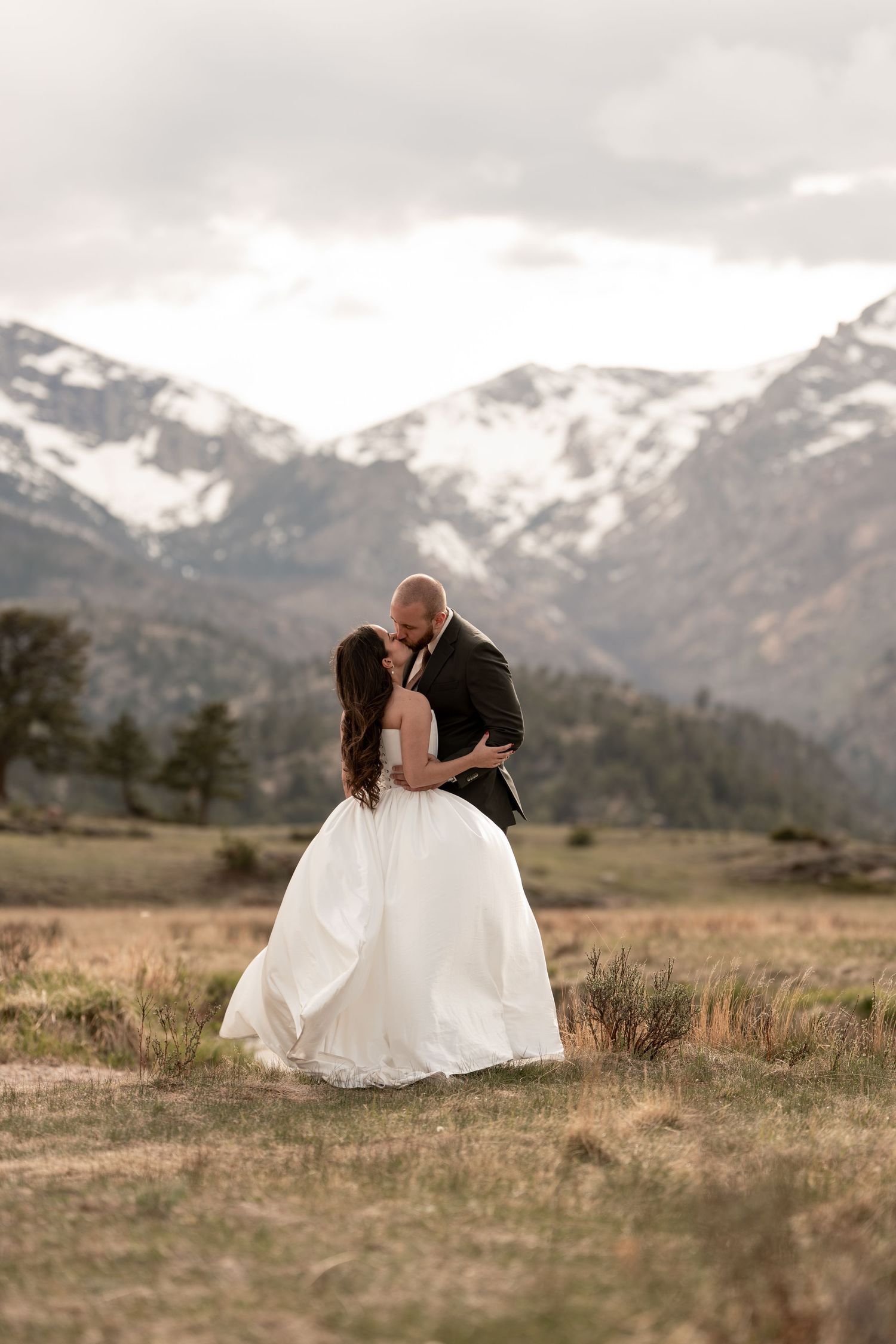 Bride and groom share a romantic kiss in a mountain meadow with snow-capped peaks in the background.