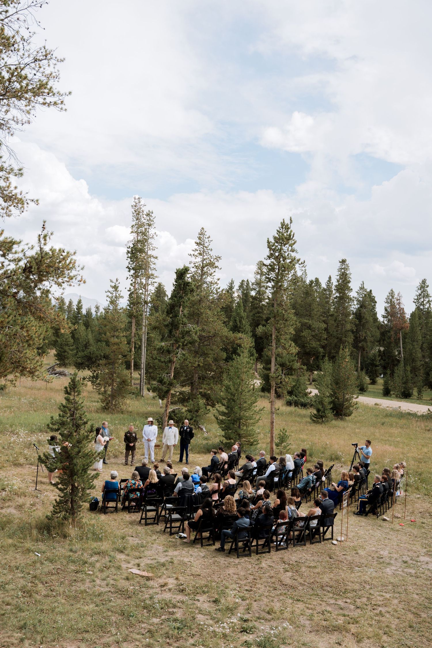 Outdoor wedding ceremony setup on a hillside with evergreen trees and guests seated in rows of white chairs.