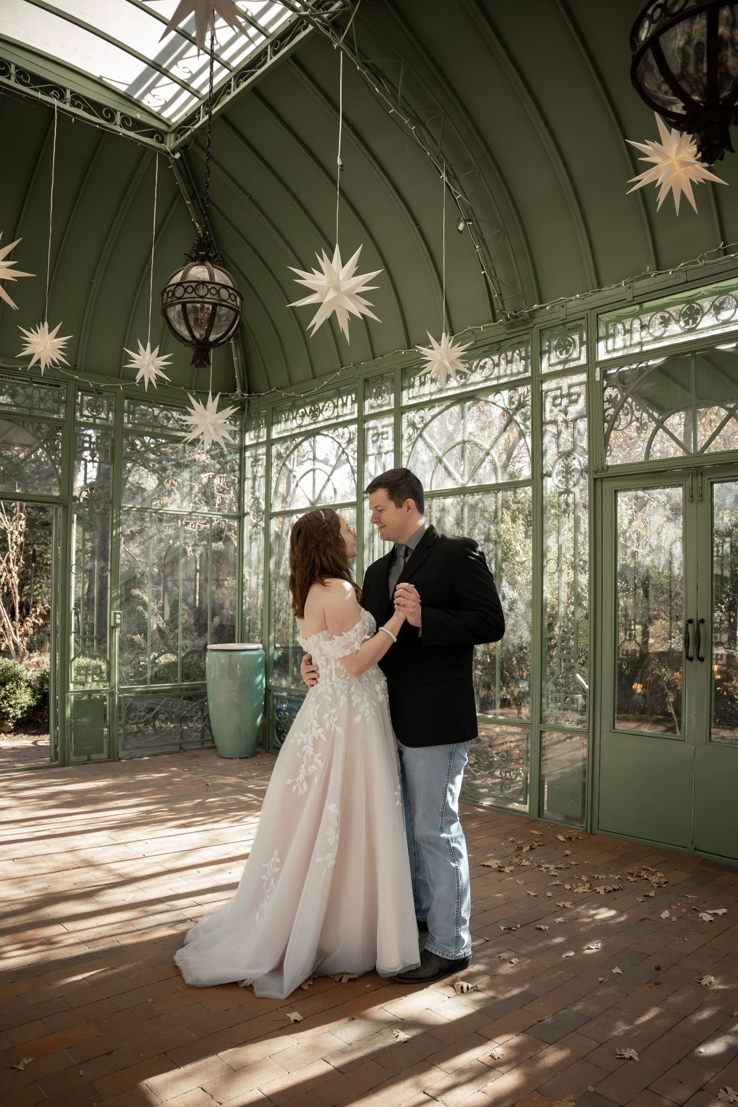 A couple dances in a sunlit greenhouse conservatory decorated with hanging star lanterns and glass windows.